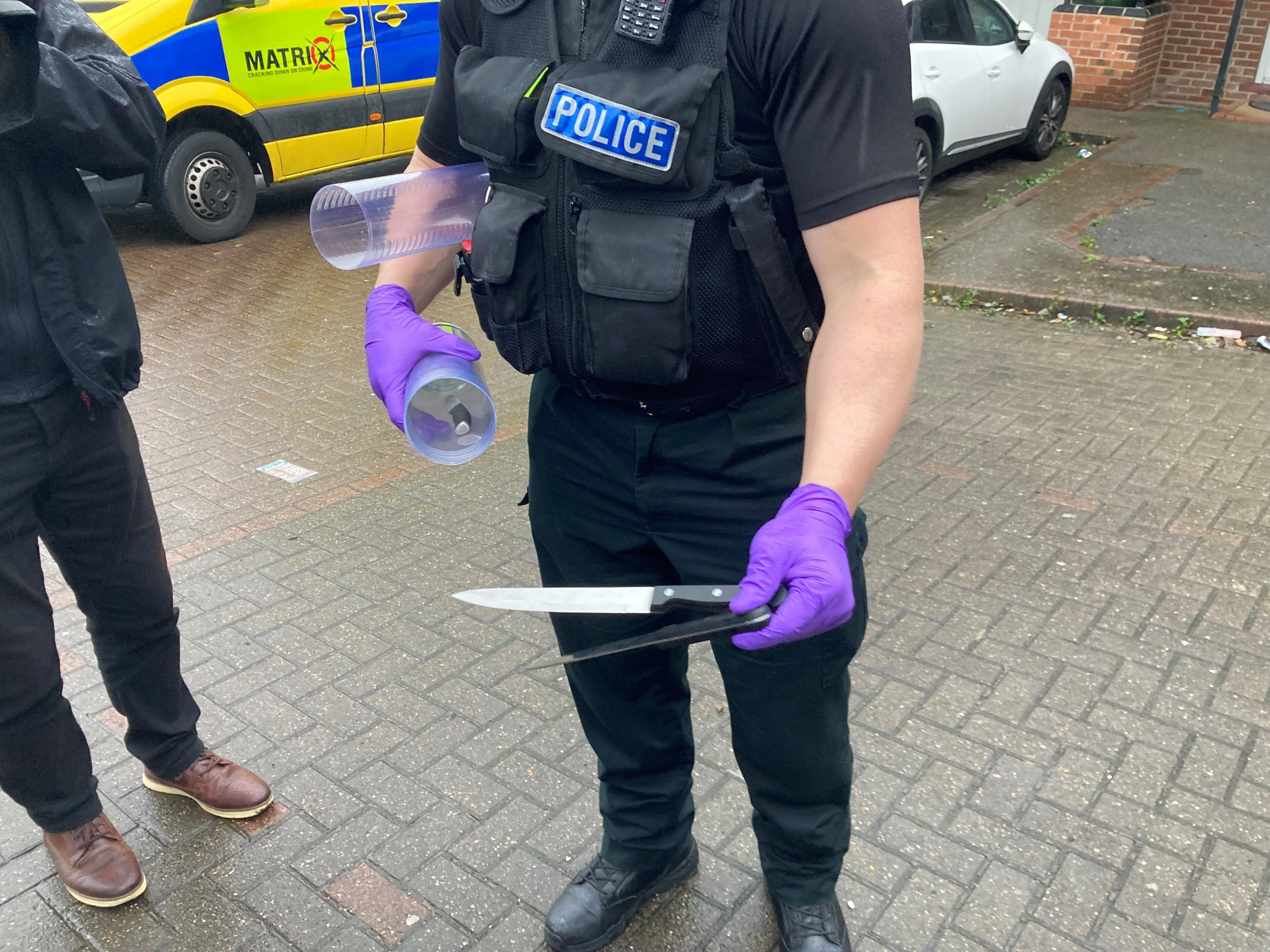 A police officer is holding up two kitchen knives to the camera. In his other hand, he is holding a plastic tube.