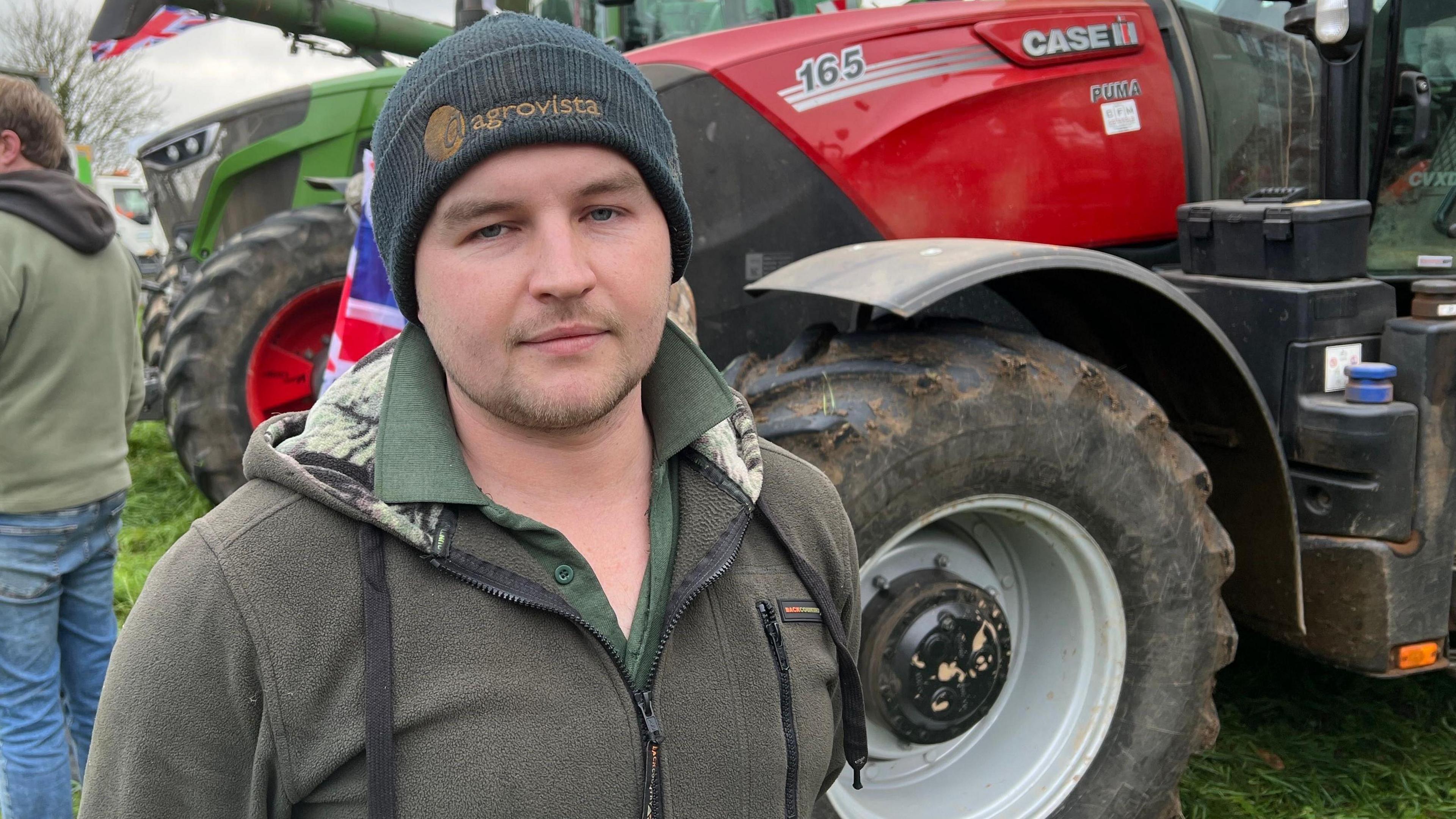 Luke Mason wearing a dark blue beanie and a grey fleece jumper over a green shirt. He is standing in front of a red tractor and looking at the camera with a disappointed facial expression. 