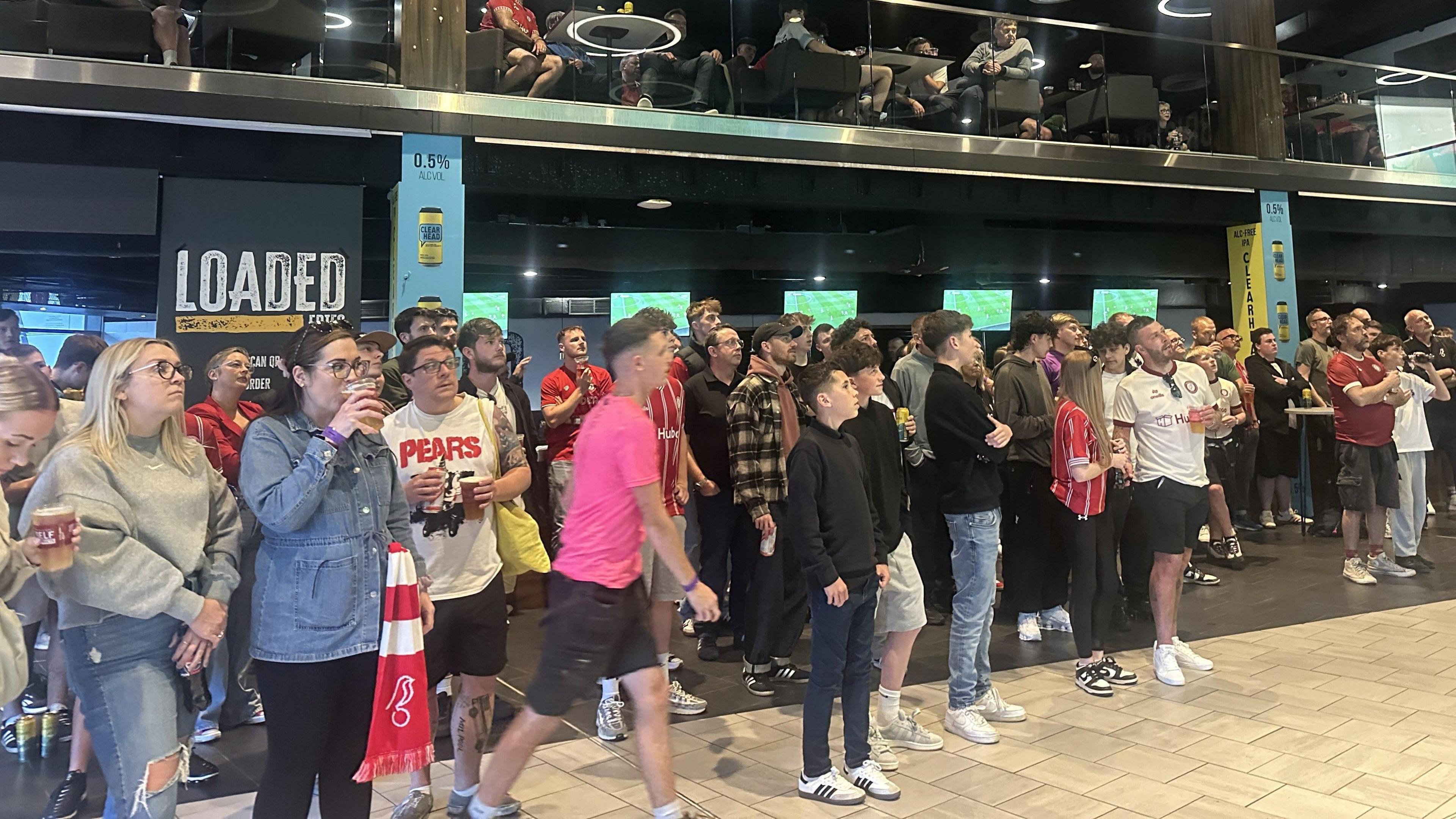 A large group of Bristol City fans, some of them holding drinks, look up at the big screen at the Sports Bar at Ashton Gate as they watch the second leg of their play-off match against Sheffield United