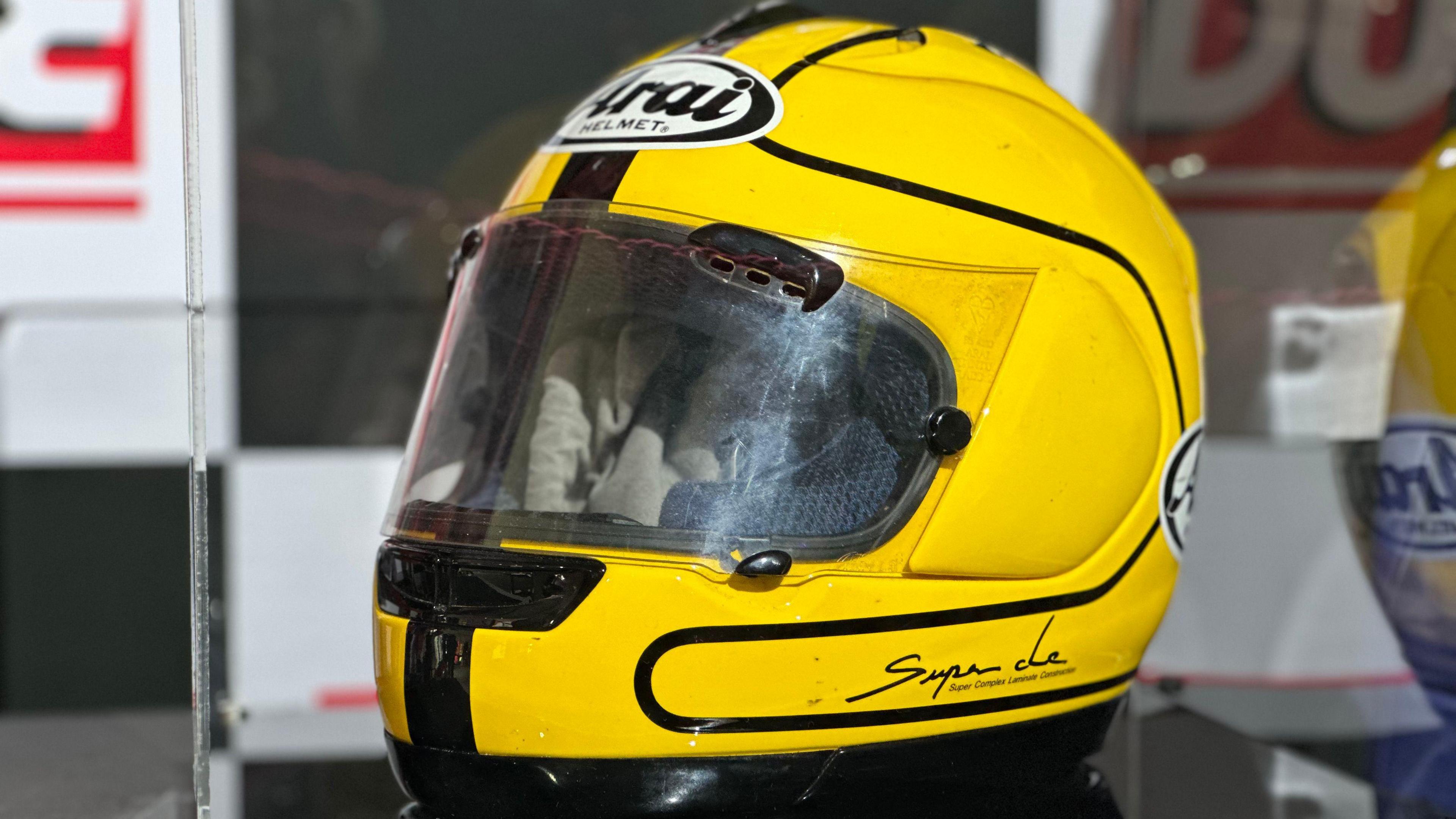 A yellow and black motorcycle helmet, with a black and white Arai logo on top. It is in a clear perspex box.