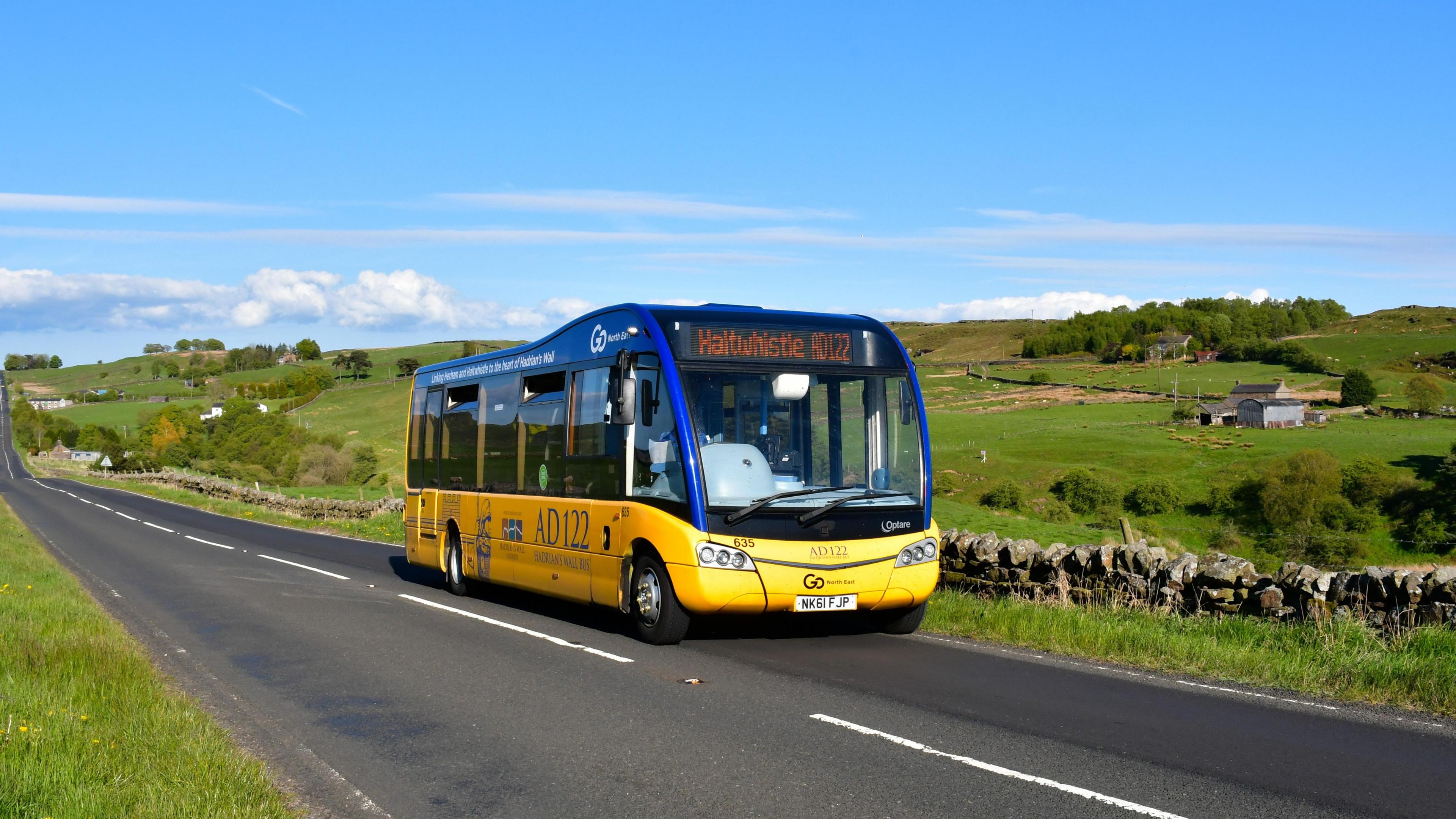 A yellow and blue bus is driving through a rural area. It has a sign on the front saying it is going to Haltwhistle 