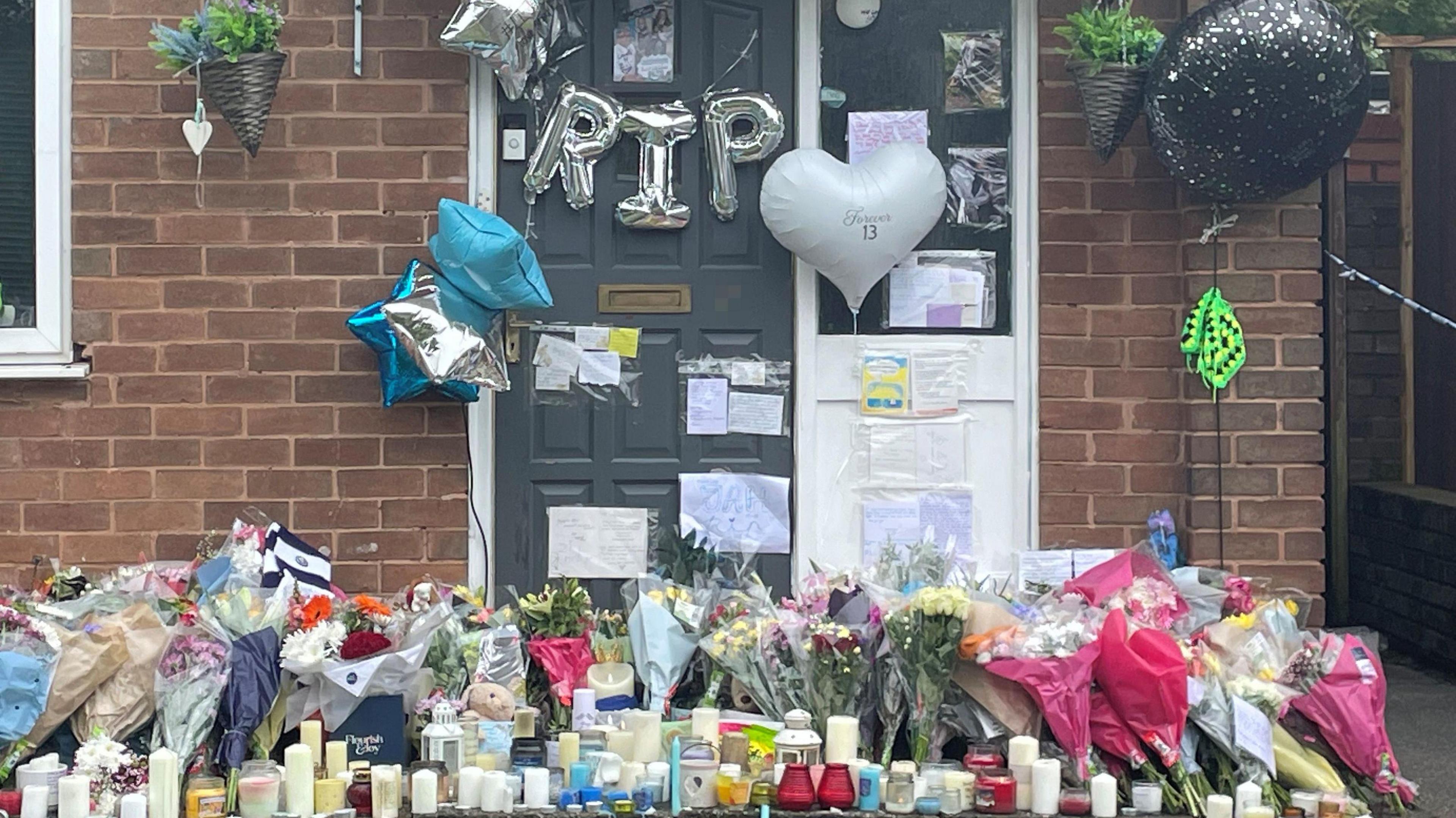 Floral tributes, balloons, candles and messages left outside a red-brick residential property with a grey front door. A number of messages are taped to the front door and silver balloons spelling out RIP hang from it.