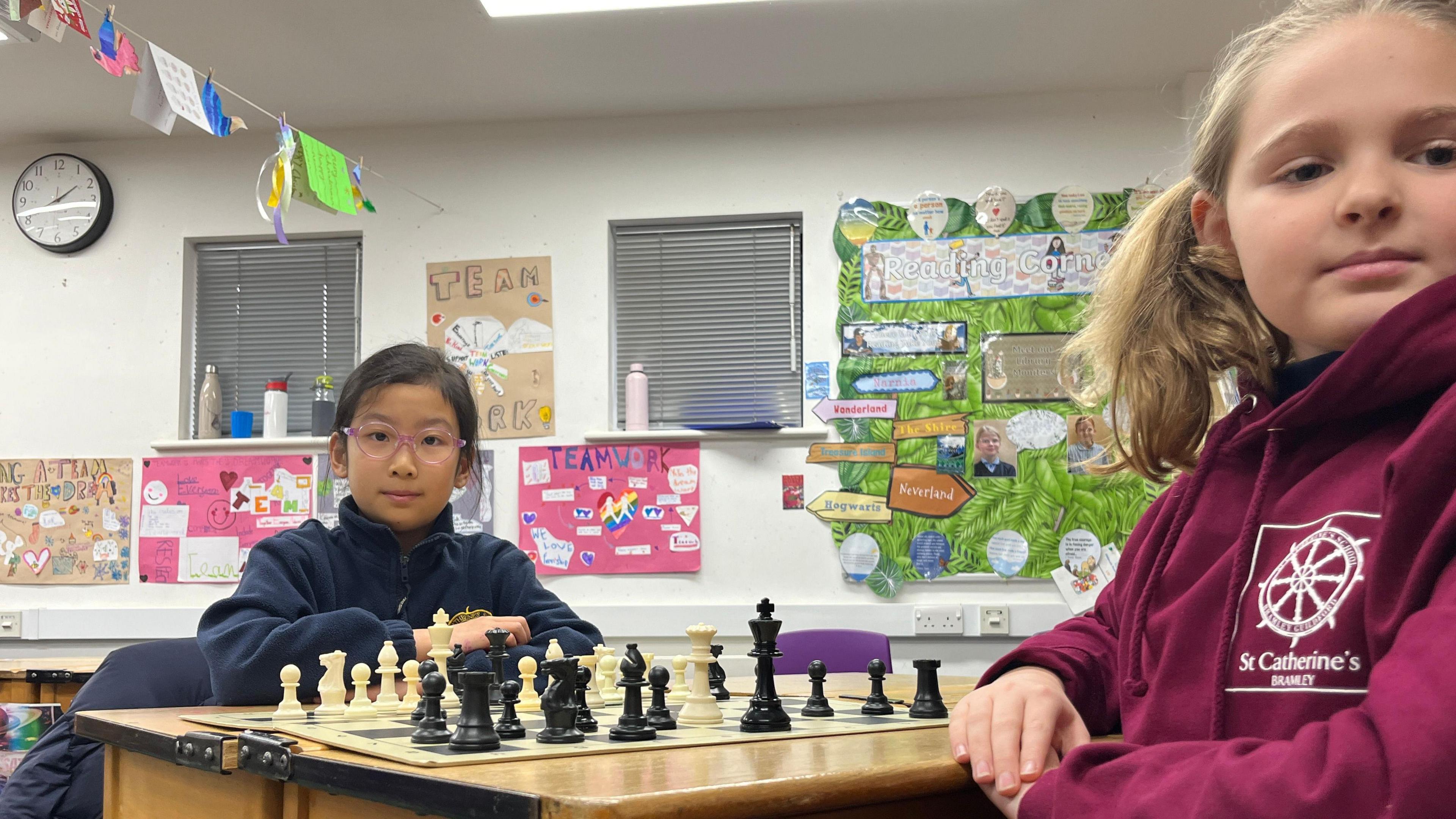 Two school girls are playing chess. One is wearing a blue jumper and one is wearing a magenta-coloured St Catherine's School jumper.