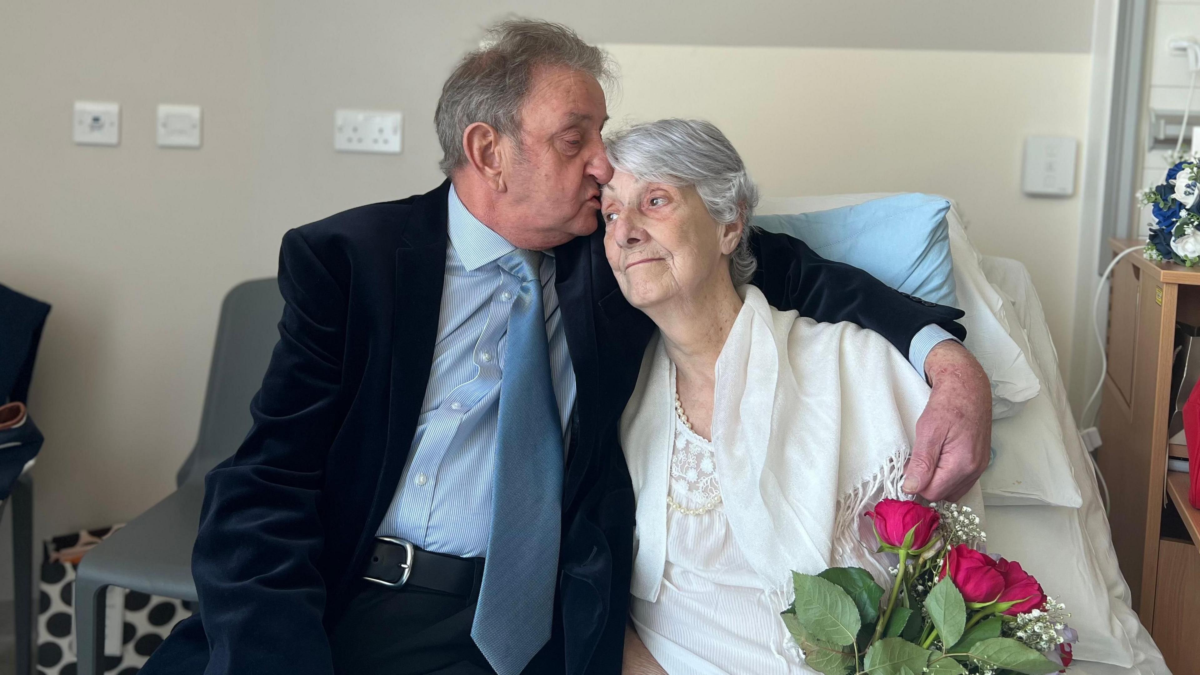 An elderly man with short grey hair, wearing a dark suit with a pale blue shirt and mid-blue tie, has his arm around an elderly woman with mid-length grey hair as he kisses the side of her head. She is wearing white. They are sitting on a hospital bed and the woman is holding a bouquet of flowers.