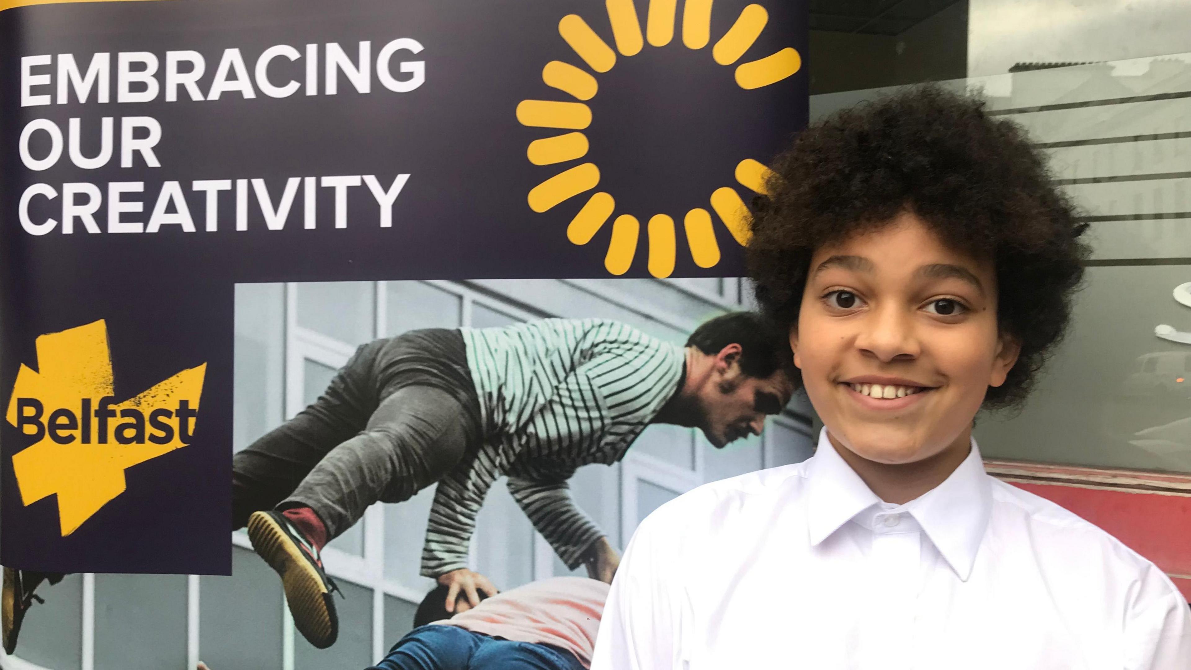 Eleven-year-old Belfast schoolboy Elliot, dressed in a white school shirt, smiling at the camera.  He is standing in front of a Belfast 2024 branded poster which says "Embracing our creativity". 