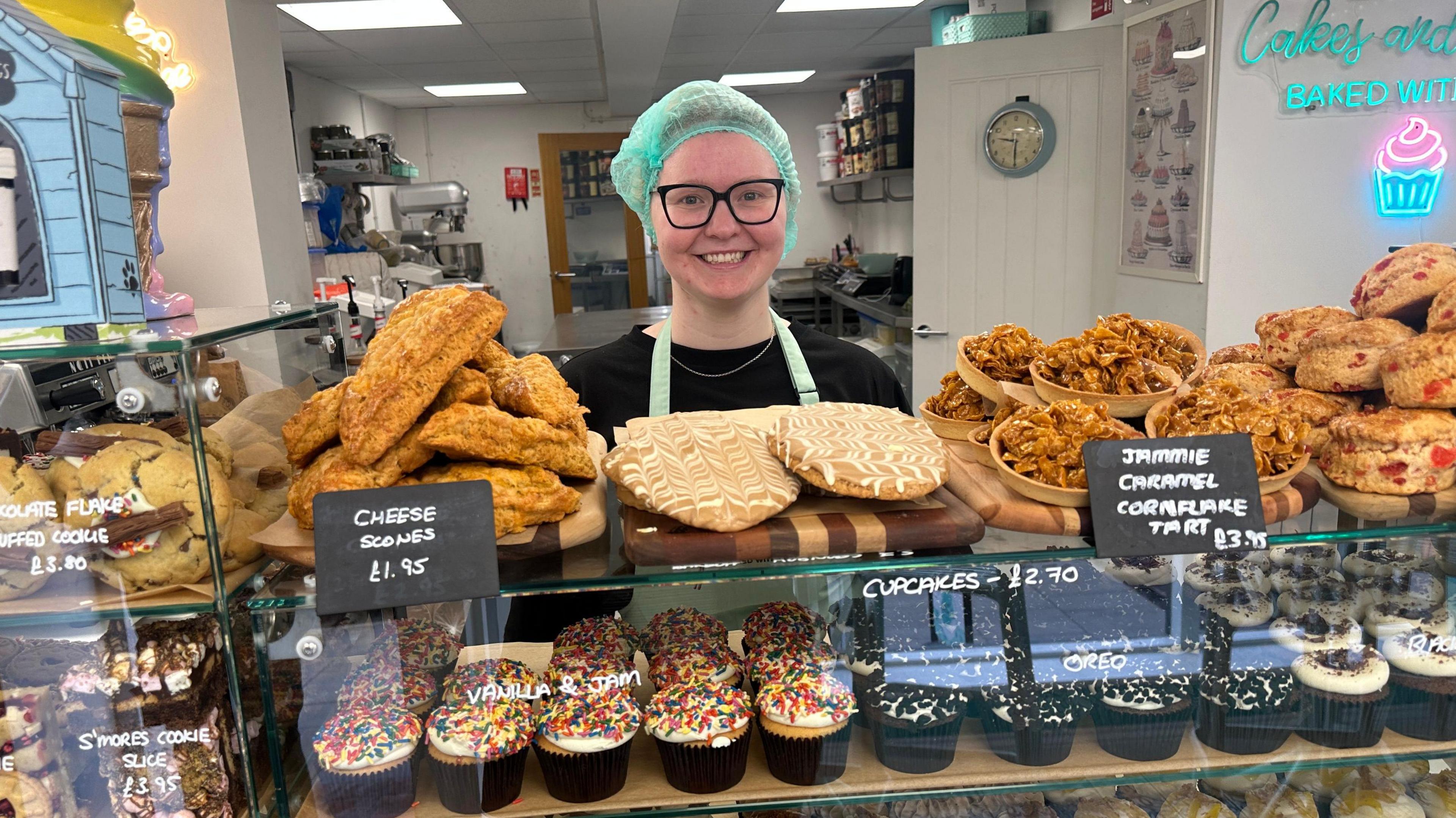 A young woman standing behind a bakery counter that is full of cakes and cookies. She is wearing a blue hair net and square glasses. 