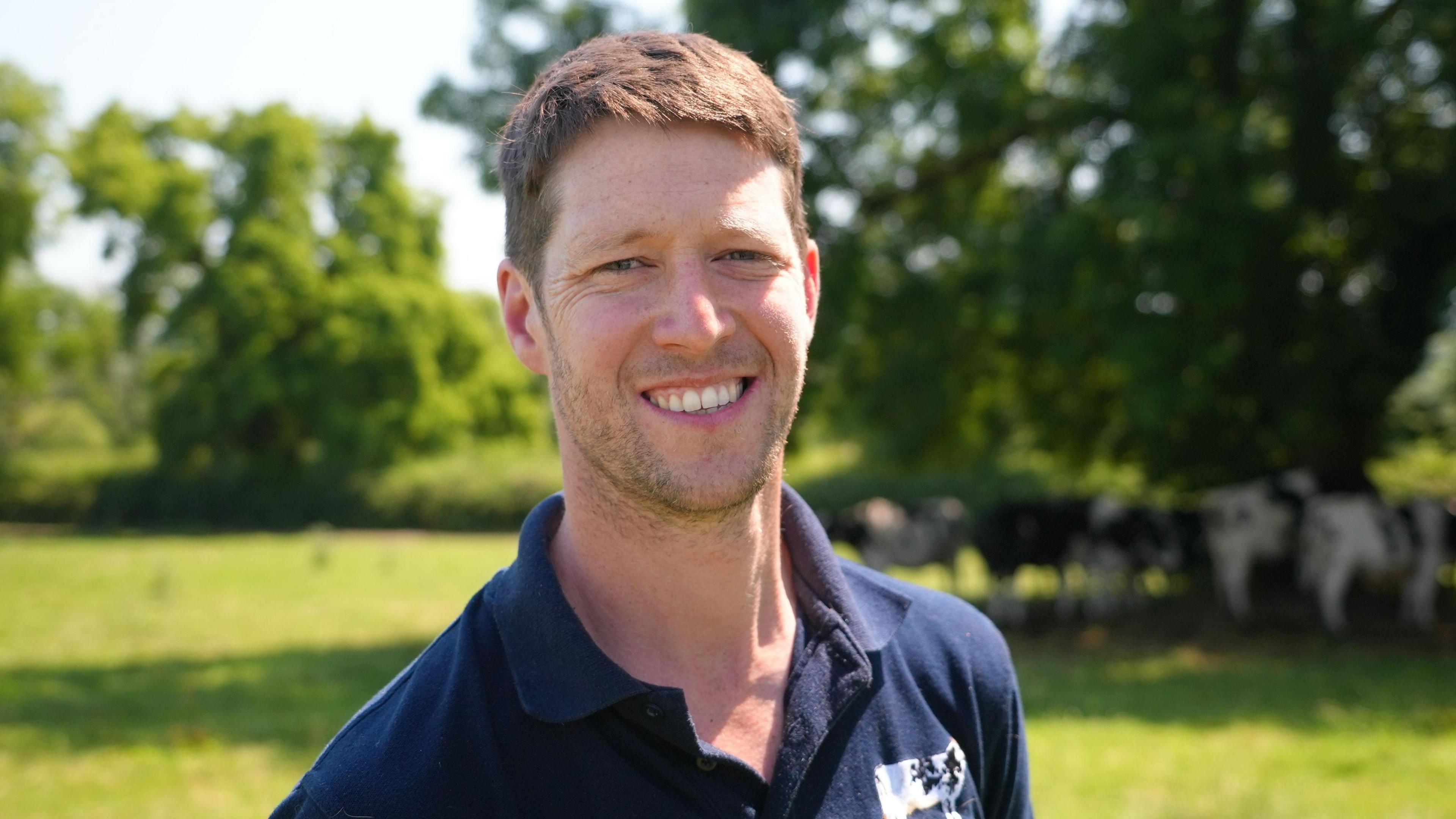 Dairy farmer Tom Jones stands in a field. He has short brown hair and is wearing a dark blue polo shirt with a black and white Holstein Friesian dairy cow logo. Behind him several cows can be seen seeking shade from the sun under a tree.
