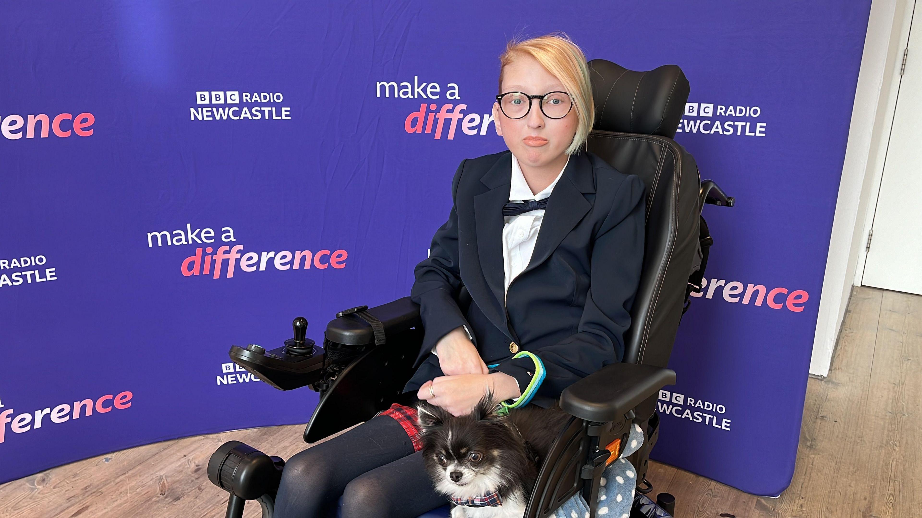 Ryoo Hooley with her dog Loki. Ryoo is wearing a tartan skirt, a suit jacket and bow tie. She is seated in her wheelchair. Loki is a chihuahua.