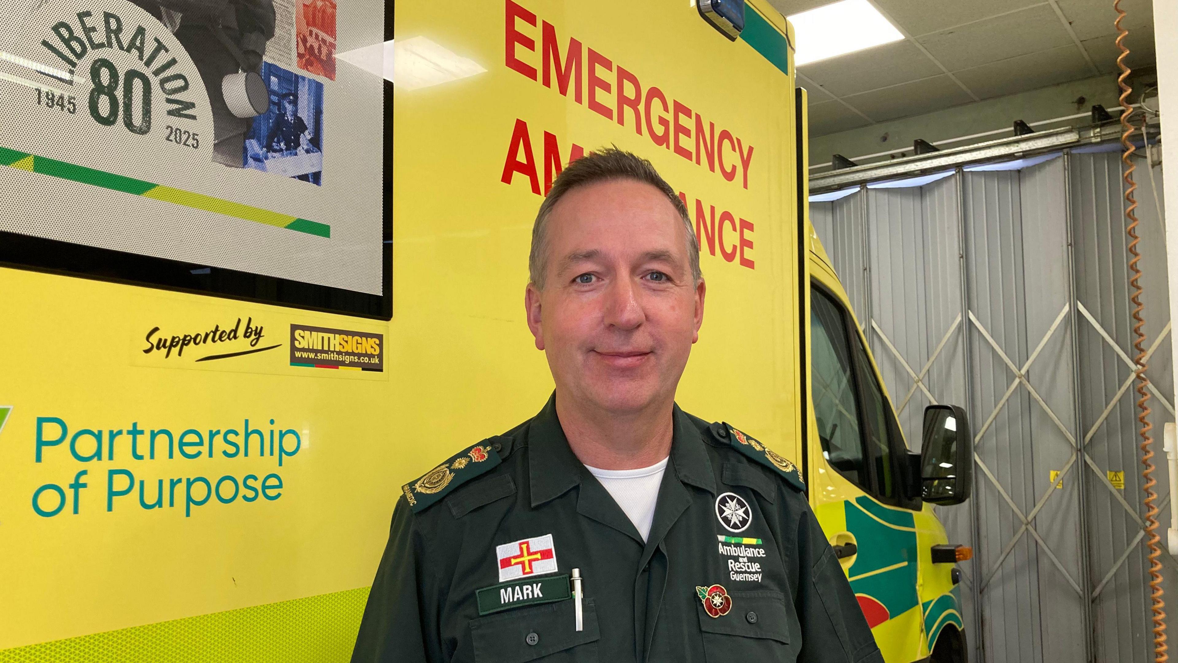 Smiling man in green uniform standing in front of yellow ambulance.