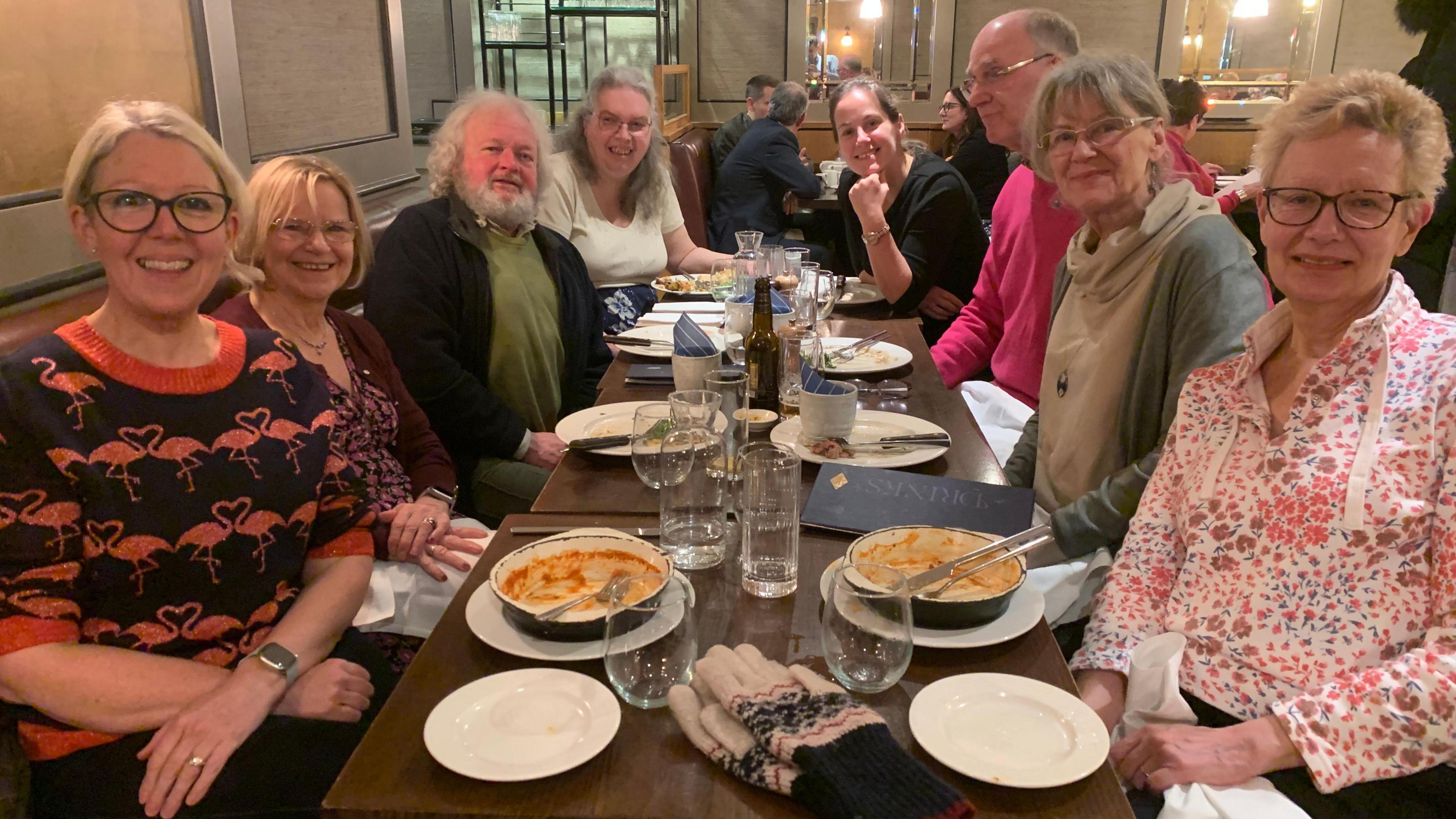 A group of eight people sat around a table, smiling as they pose for a photograph at a restaurant. They are all aged middle aged or older.