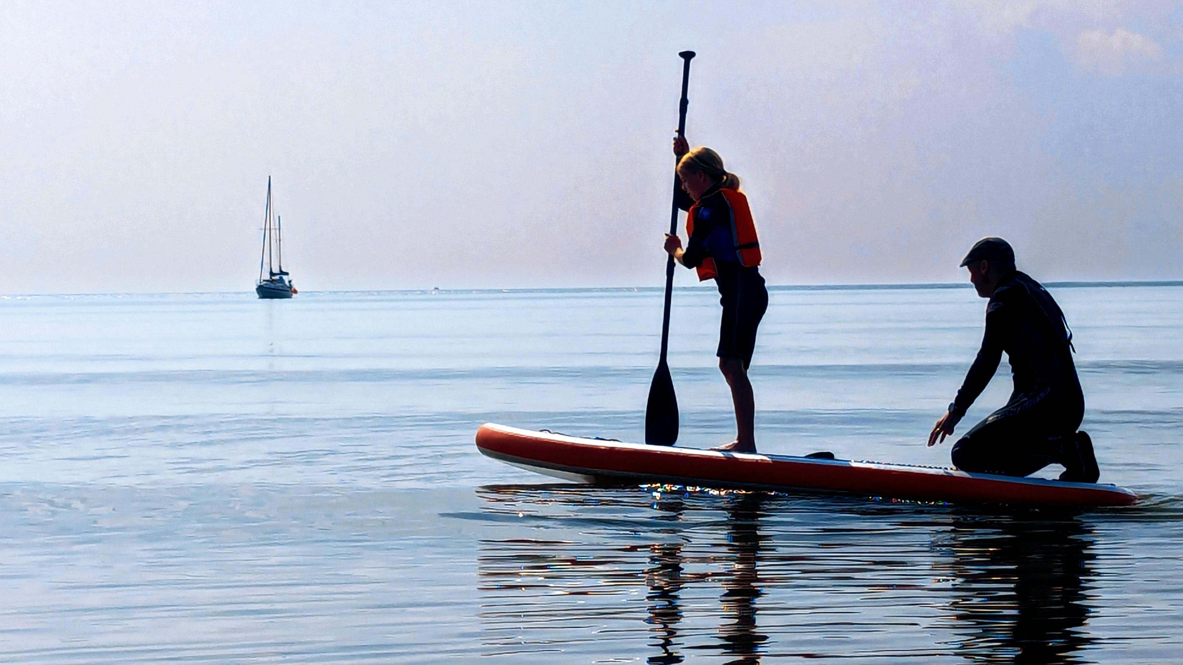 Two paddleboarders at Brigg Bay. A young girl in life jacket is standing on the board while a man wearing a hat is sitting behind her. There is a boat in the background.