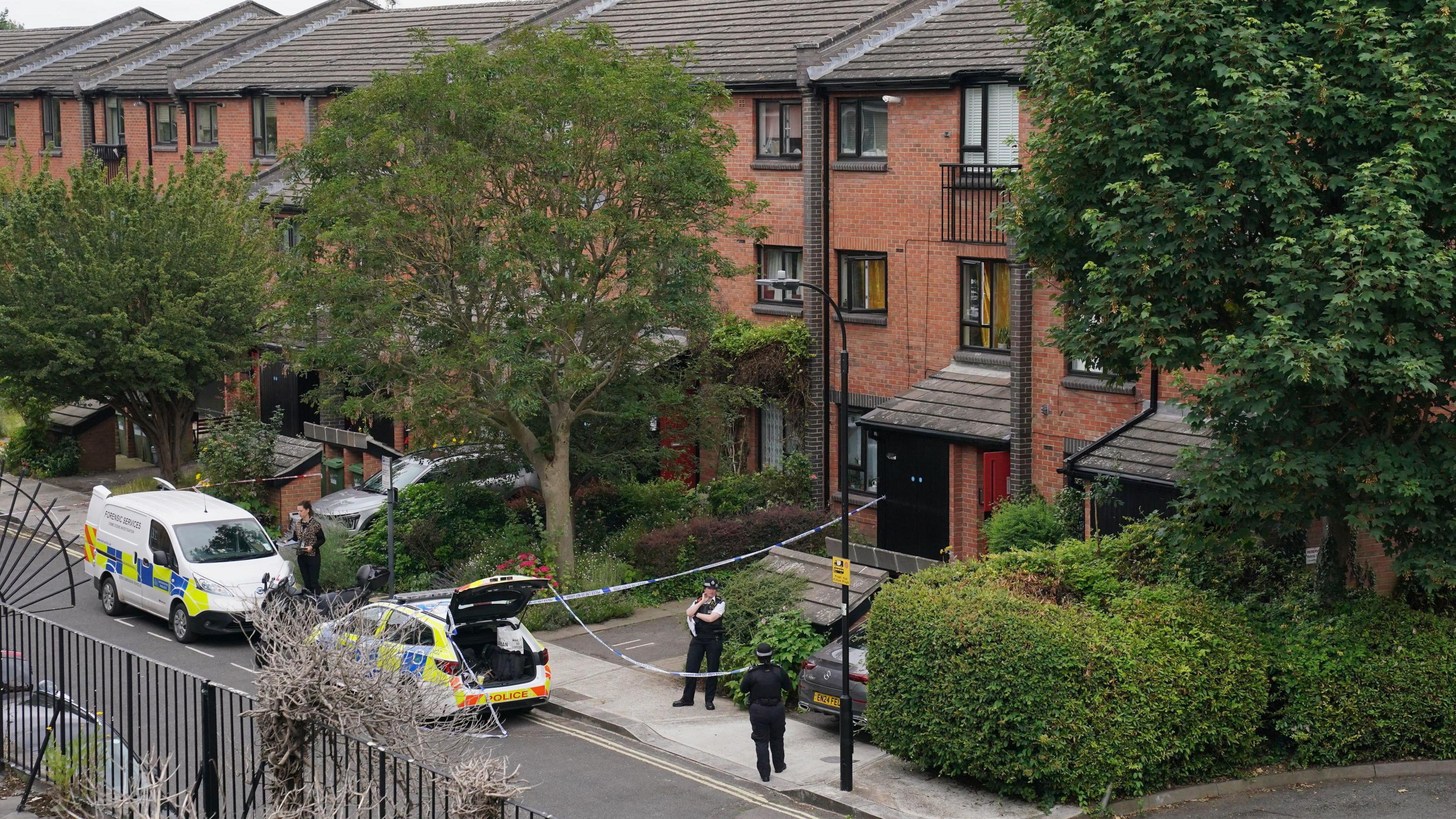 Two police officers are standing outside Paul and Albert's flat in 9 Scotts Road, London. There is a police cordon around it, with a police car's boot open and a forensic van in front of it.