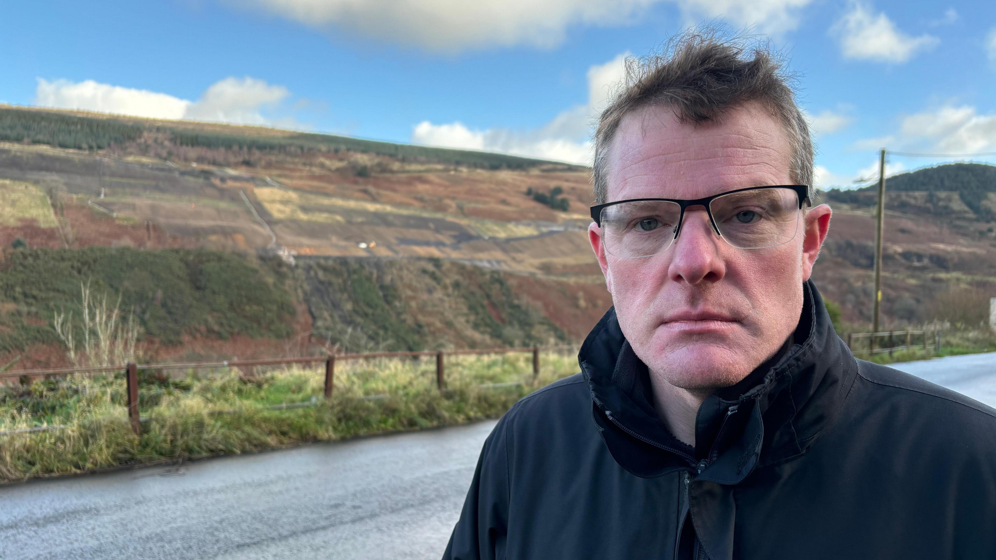 Phil Thomas, with short, dark hair and wearing glasses and a black coat, is to the right of frame. Behind him is a road and in the distance is a hillside which is the site of a coal tip which has been made safe. Aside from the green, yellow and red colours of the grass and bracken on the hillside, there are also patches of black which is the coal tip.