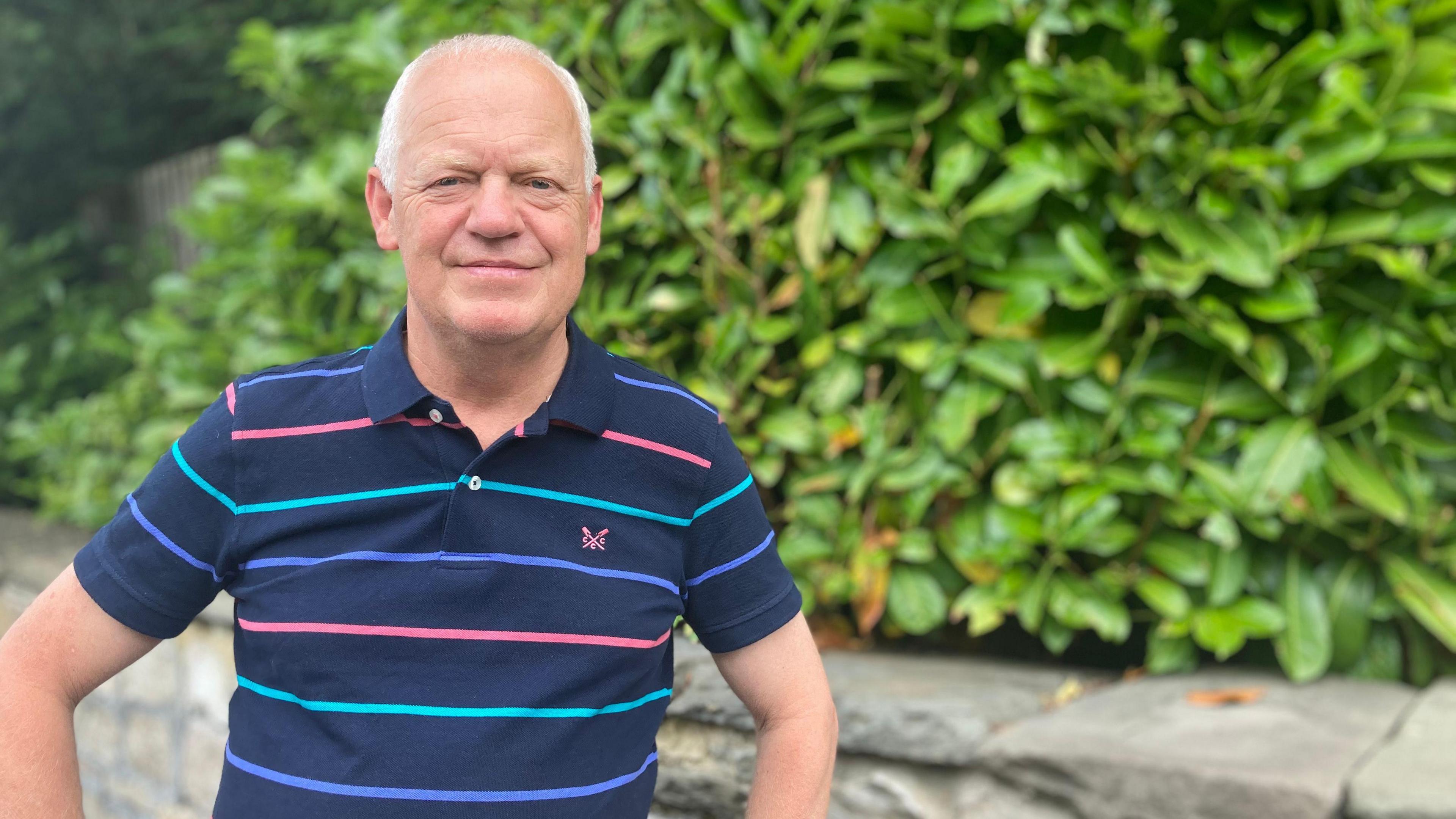 Paul Atwell in a stripped blue t-shirt standing in front of bushes and a wall.