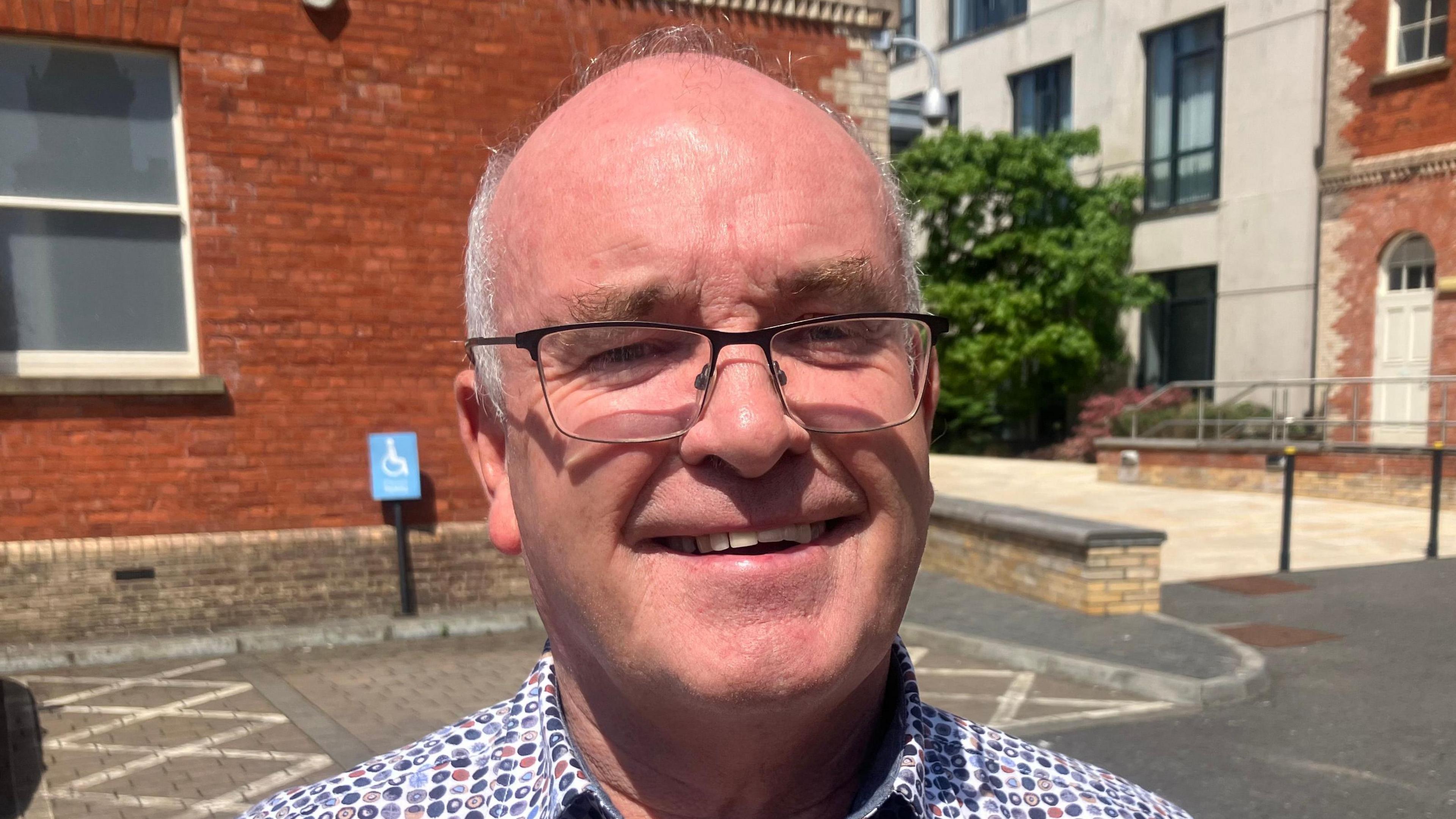 A balding man with rimmed glasses and a spotty shirt smiling in front of a car park in front of red brick buildings.