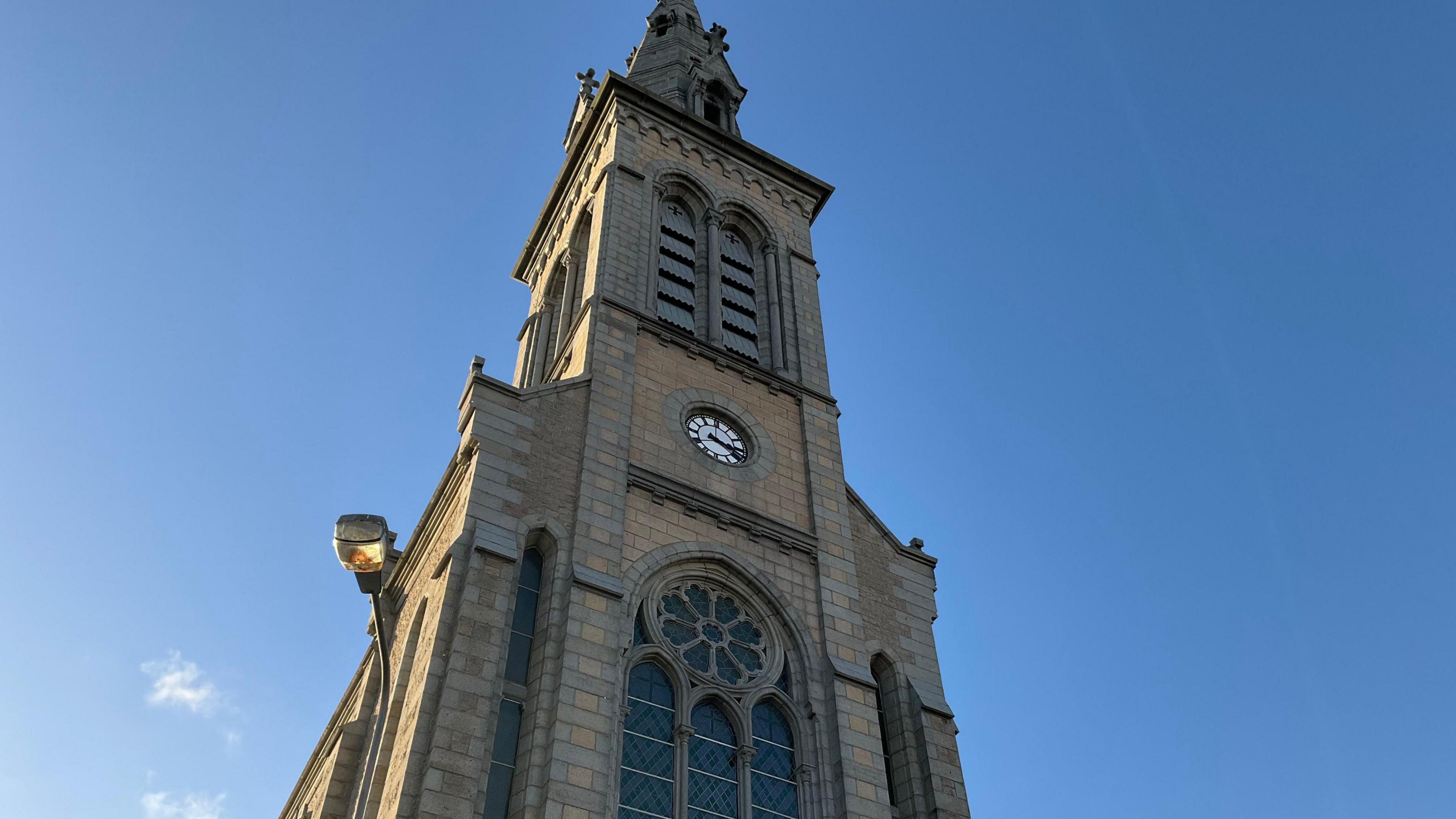 The outside of a church tower on a clear sunny day. The camera is looking up at the church clock tower. There is clear blue skies with only a few clouds in the sky. The church has a clock and some large windows.
