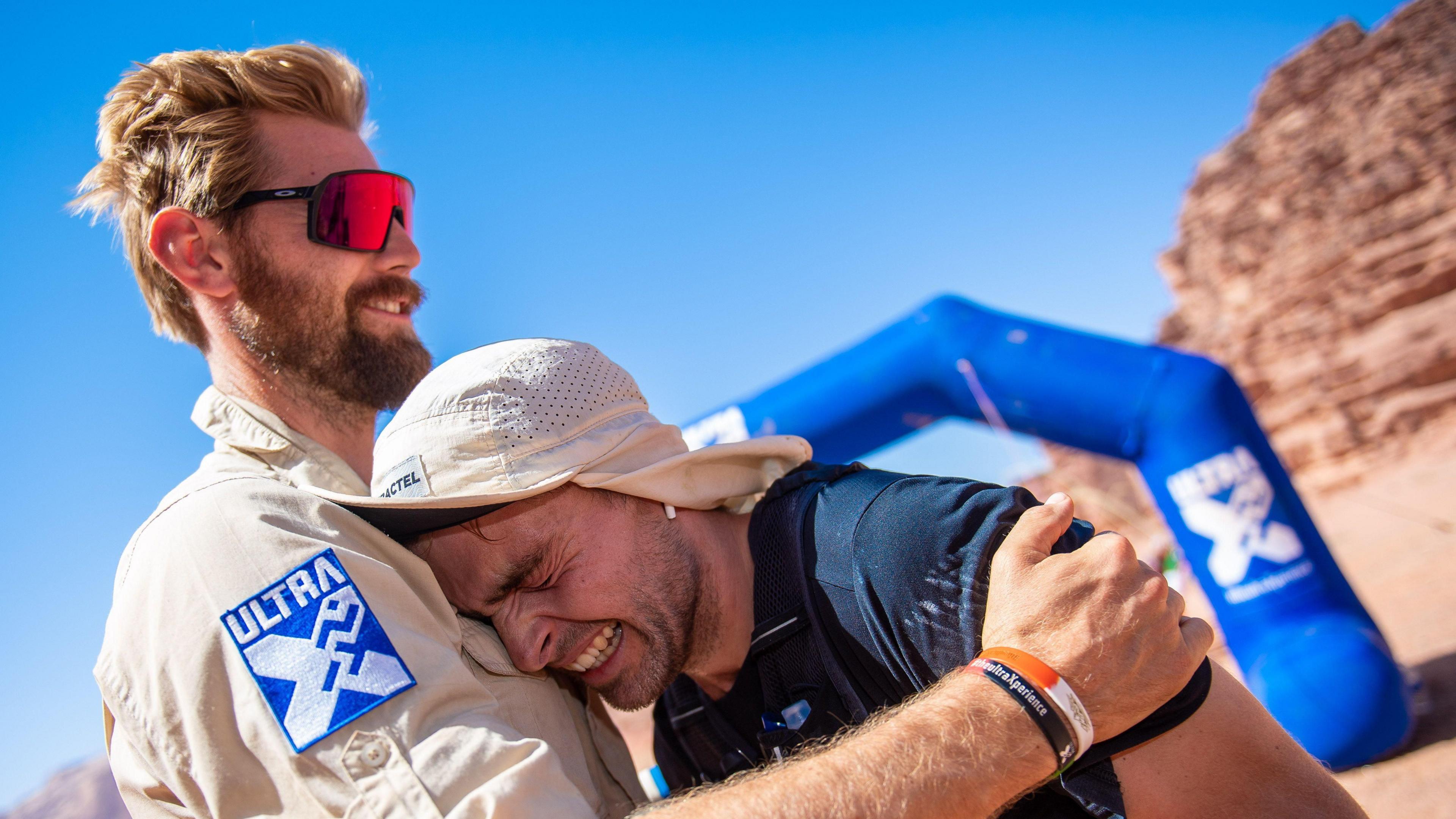 Sam King crying into the chest of a man having completed an ultra marathon. The man has a cream shirt on and sunglasses. Sam has a cream hat on and a navy T-shirt.