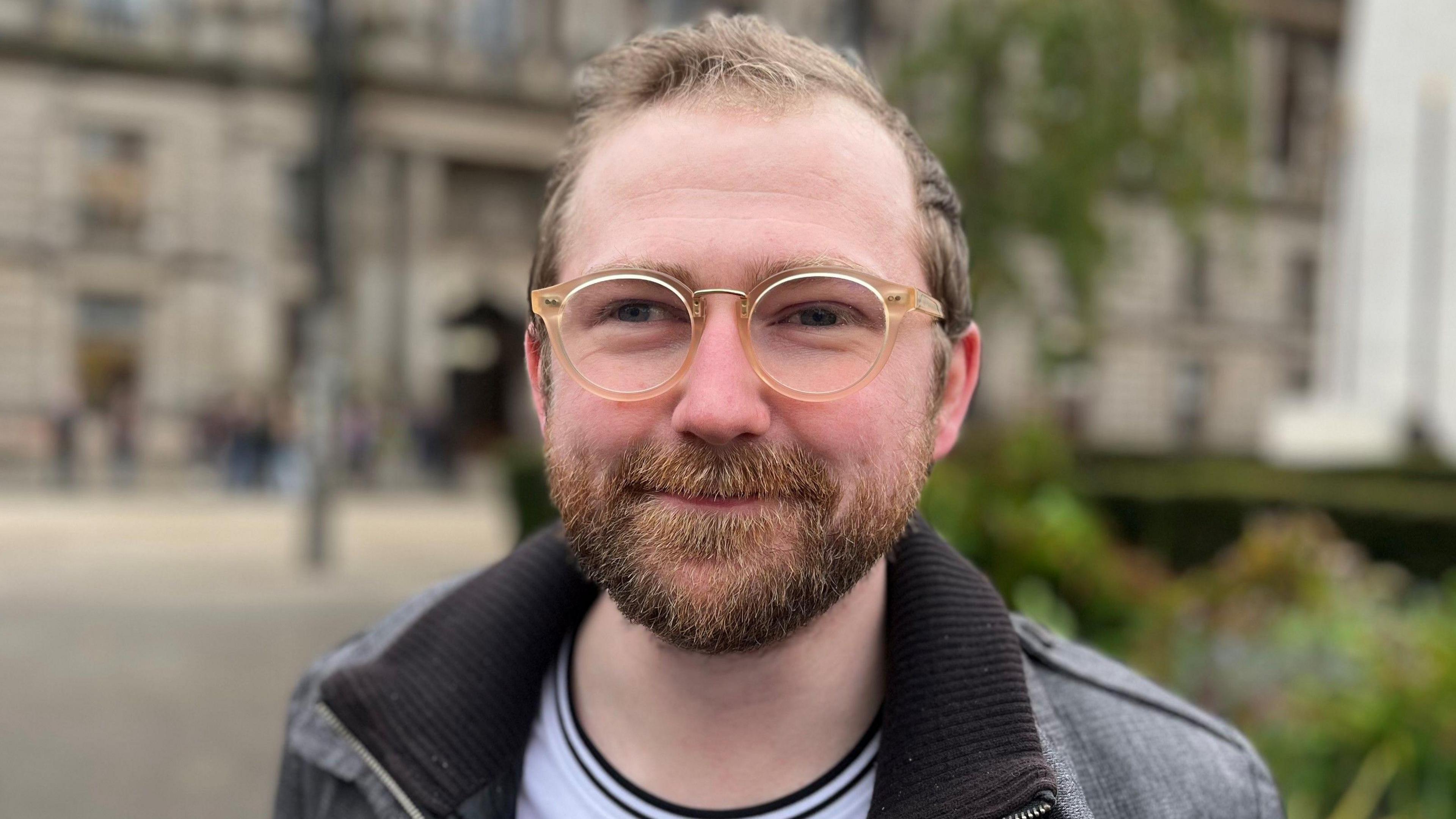 man with beard and transparent framed glasses head and shoulders shot looking at camera. Blurred indistinct building in background