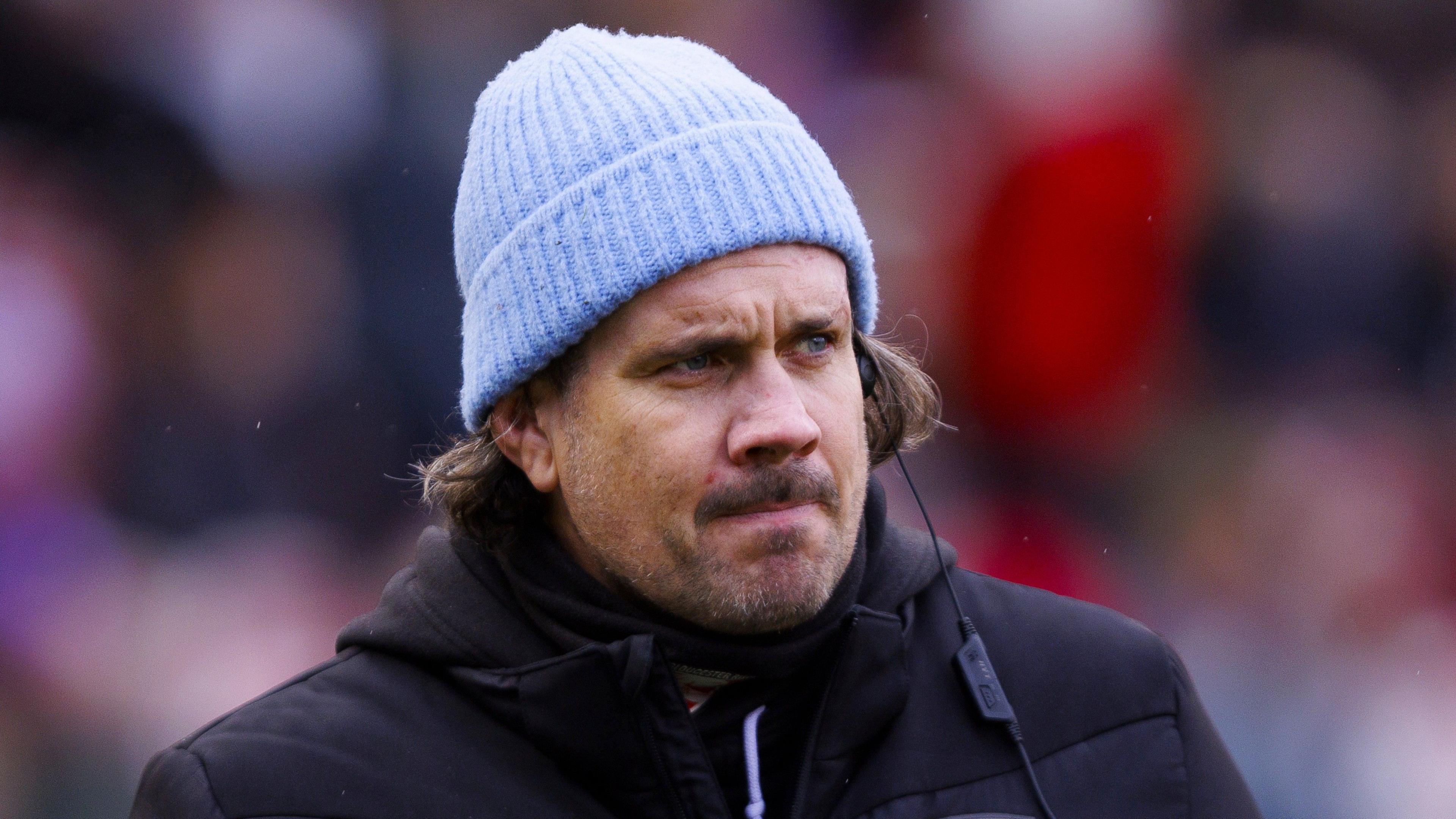 Gloucester-Hartpury head coach Dan Murphy looks on while wearing a black jacket and a blue hat before a Premiership Women's Rugby match.