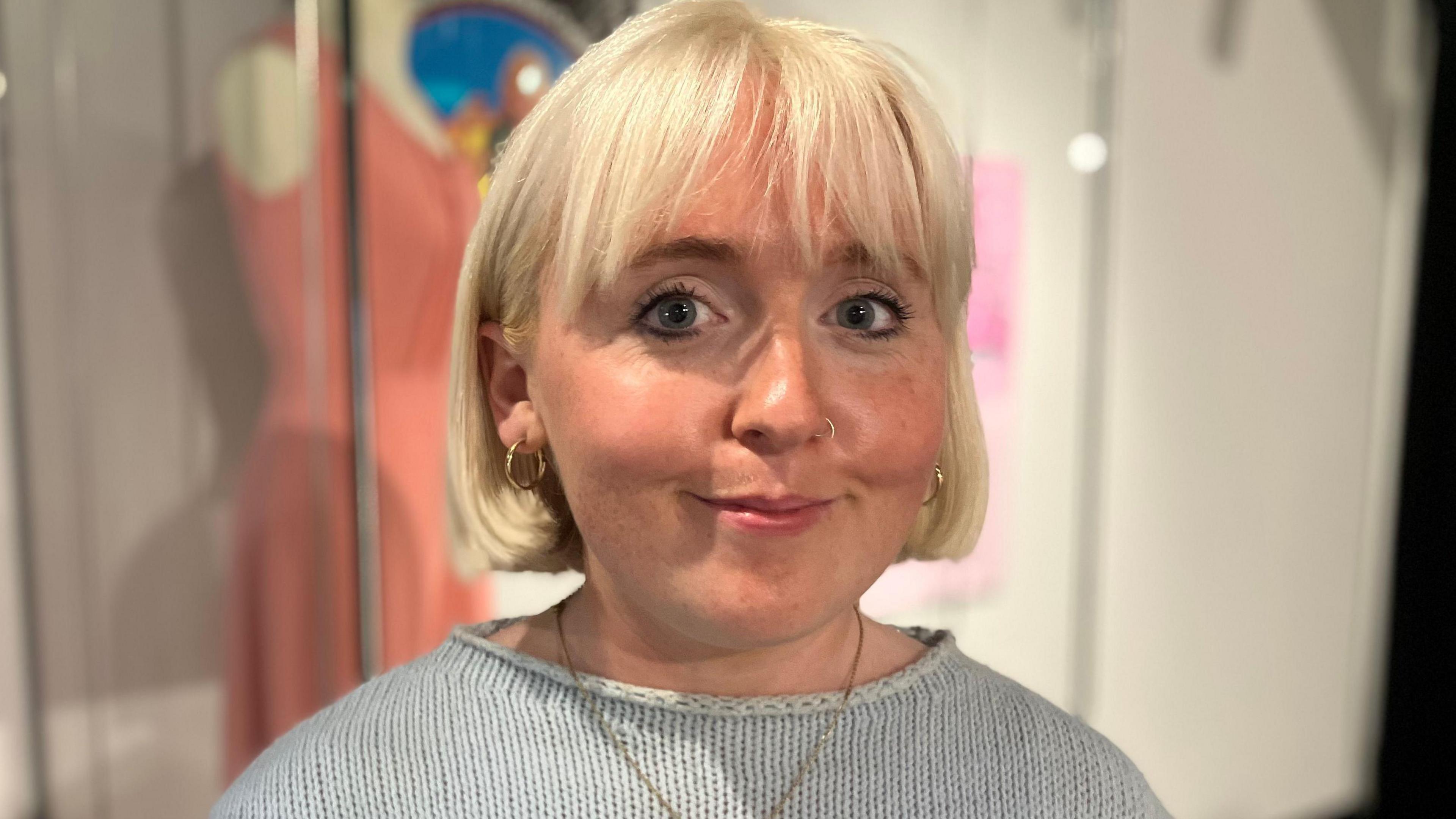 A woman with a white blonde bob looks at the camera. She wears a baby blue jumper with a gold chain hanging from her neck. Behind her, a "coal queen" dress is blurred.