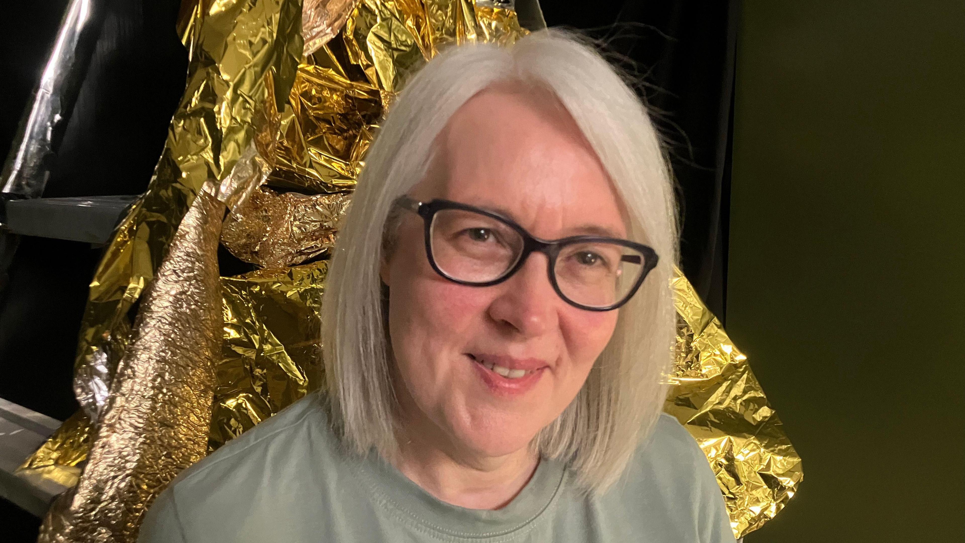 A woman, pictured from the shoulders up, smiling at the camera. She is wearing a green top and glasses and she has blonde hair.