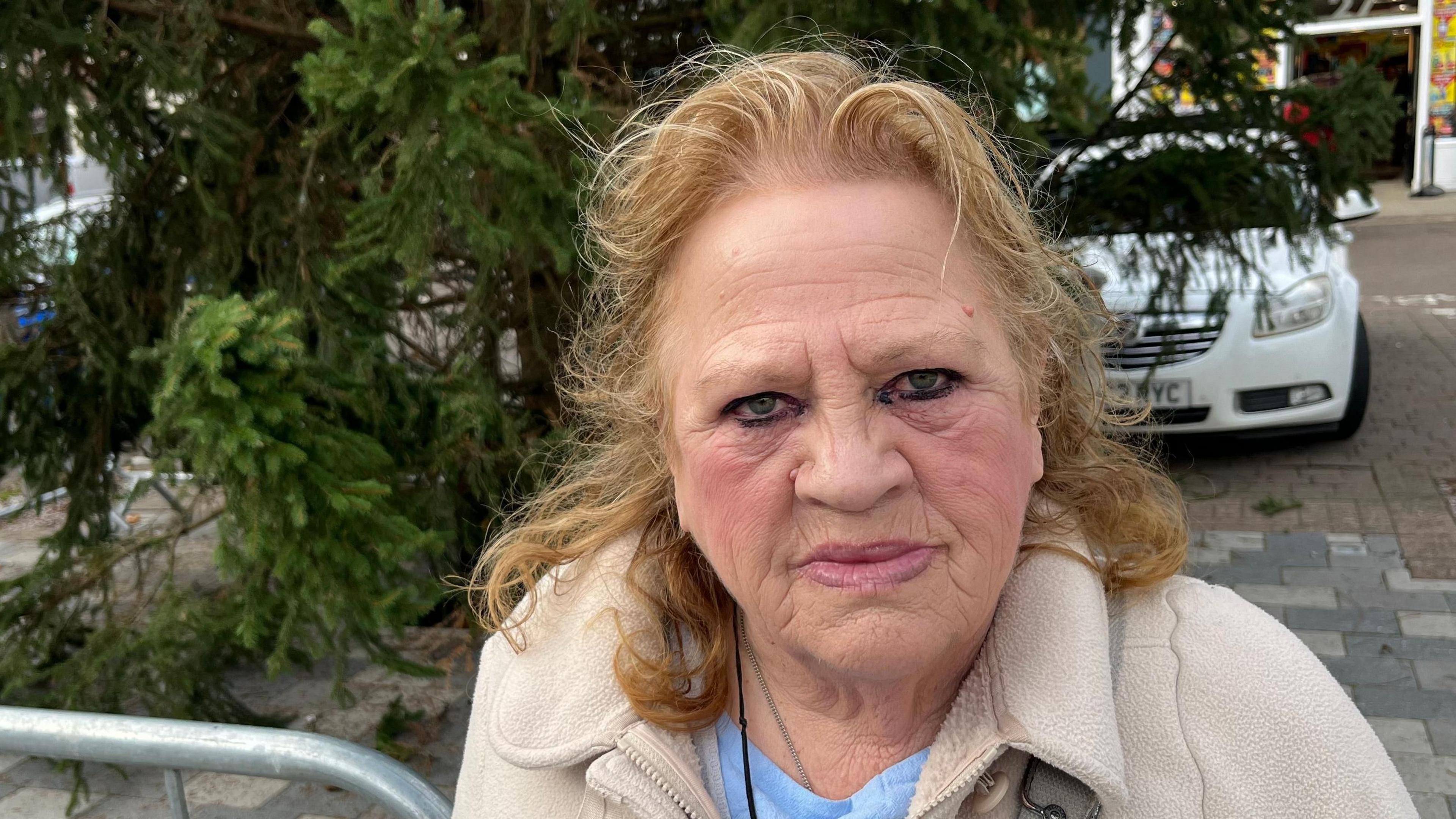 A woman with strawberry blonde hair, a beige fleece and blue top looks at the camera. Behind her are the lower branches of a Christmas tree placed on grey paving slabs