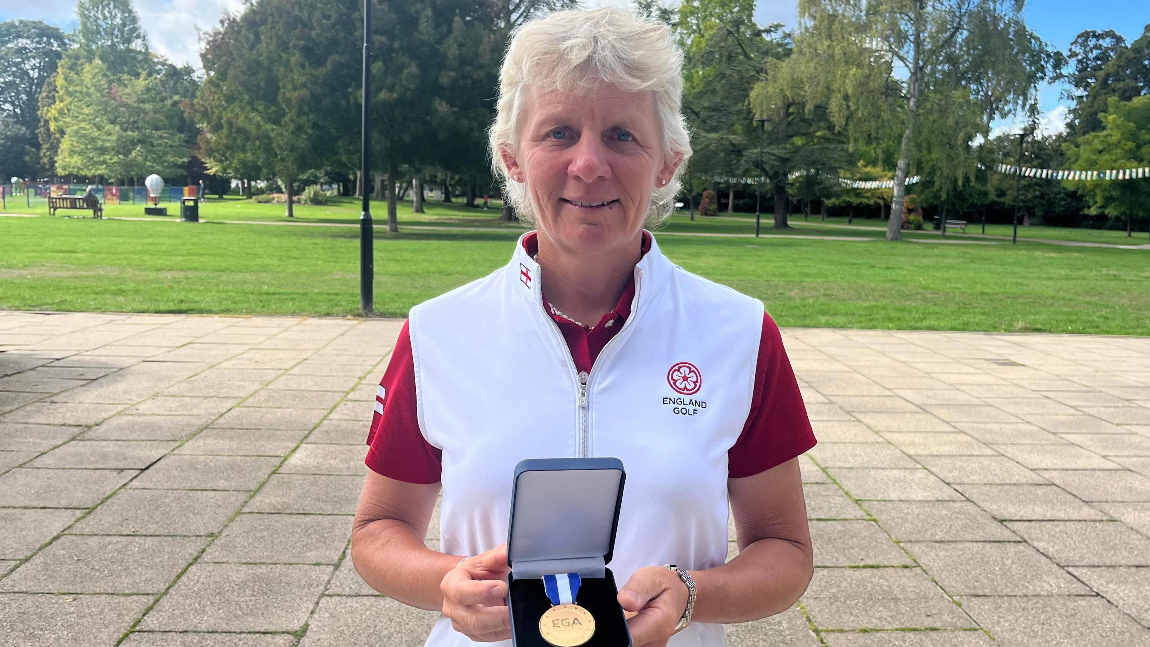 Sally Shayler in her red and white branded golf attire, holding her medal in its casing with both hands. Behind her is a park, and she is standing on paving.