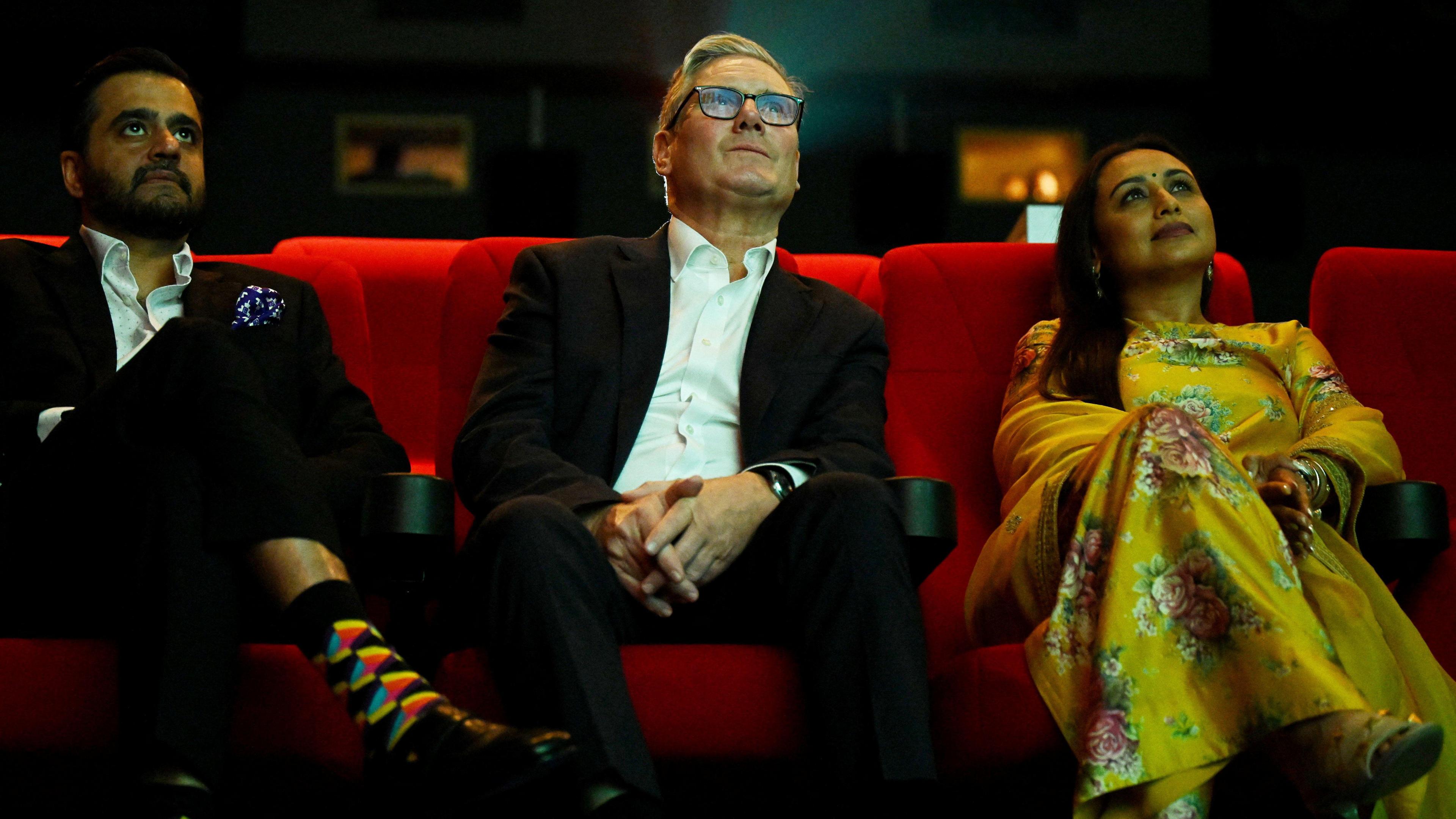 In a dimly lit movie theatre, sit Yash Raj Films CEO Akshaye Widhani in a dark suit, white shirt, and colourful socks; British Prime Minister Keir Starmer wearing glasses and a dark suit with a white shirt; and actress Rani Mukerji in a yellow floral outfit. They are seated on red theatre chairs, facing the screen.