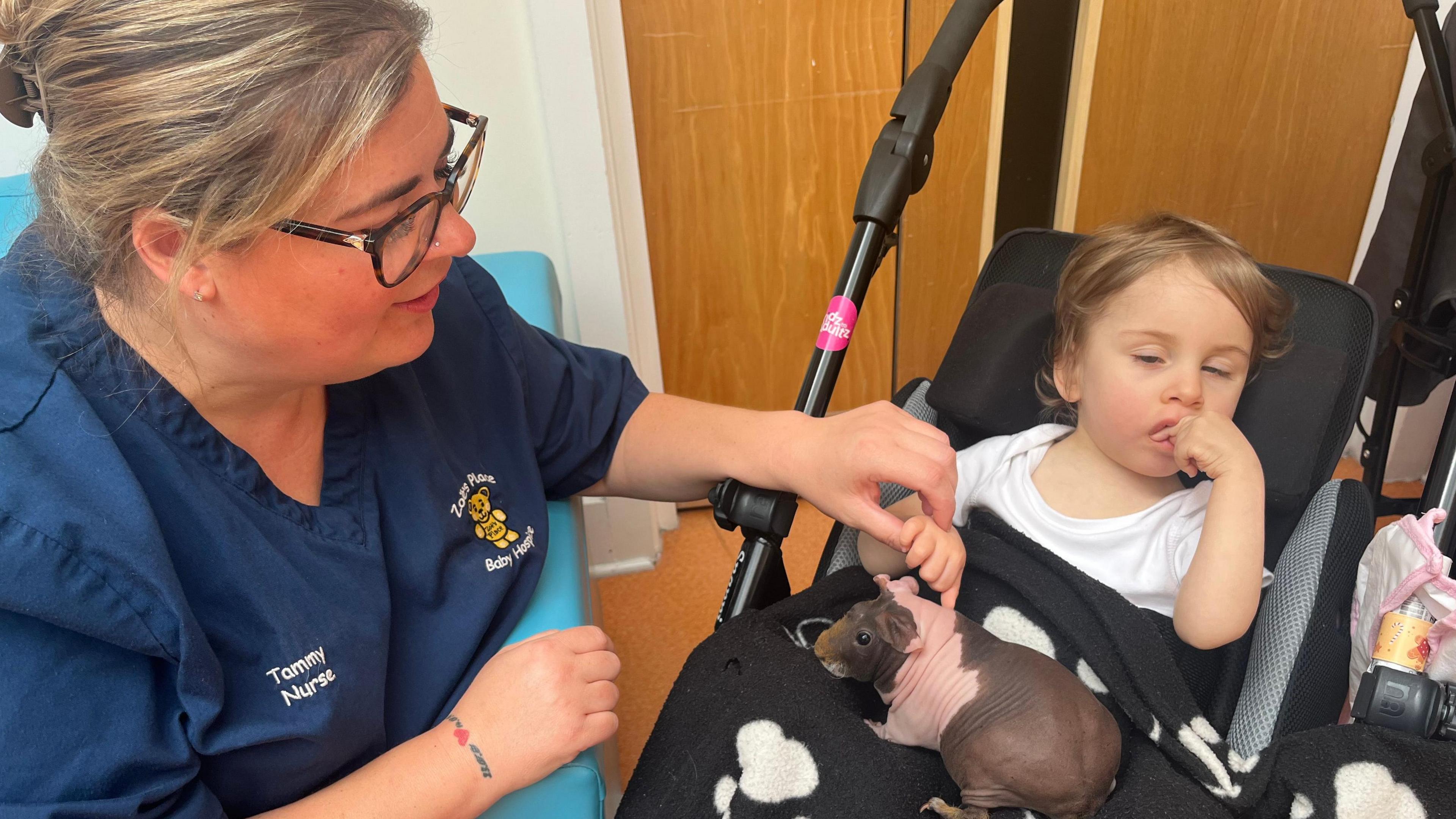 A blonde nurse in blue scrubs with a teddy bear logo on, sitting next to a young girl with brown hair in a pram. The girl has a black blanket with white paw prints over her lap, with a hairless brown and pink guinea pig on her lap.
