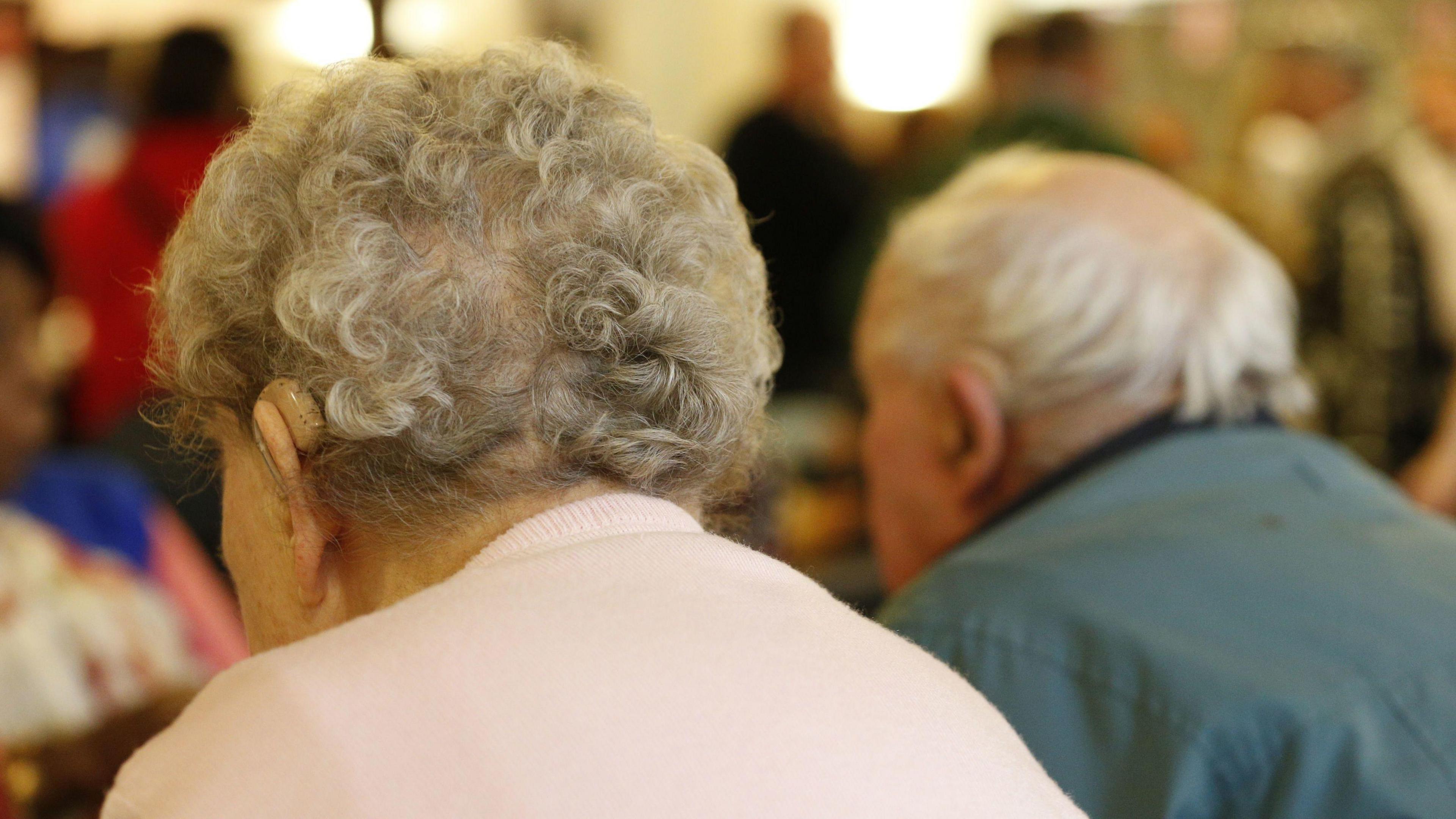A stock image of two older people, sat with their backs to the camera. An elderly woman is in the foreground of the image. She has short grey curly hair and wears a pale pink jumper and hearing aid. An older man sits in the background. He has grey hair and wears a green jacket, as well as a hearing aid.