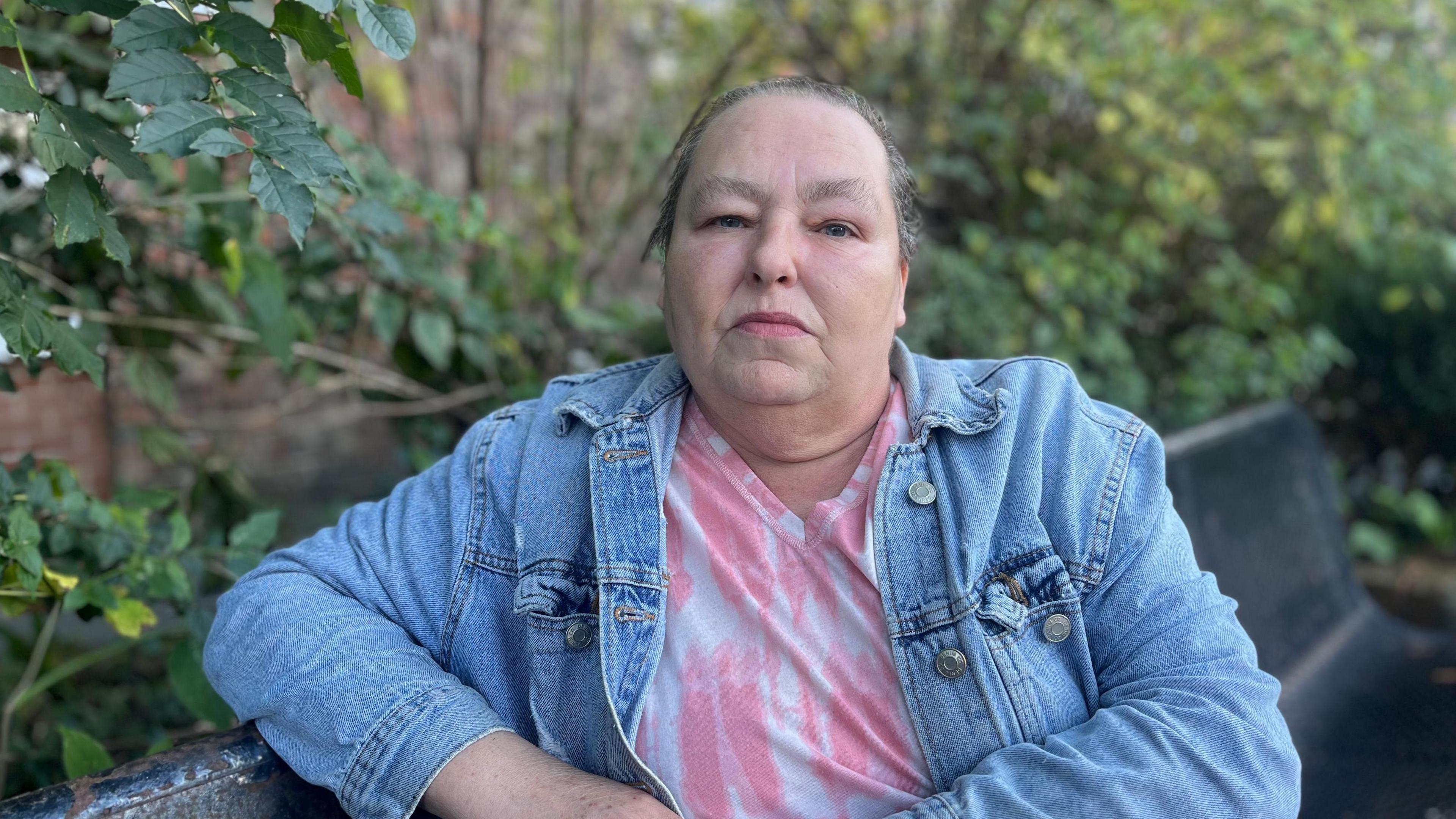 Elizabeth Neailey is staring into the camera. She's got dark hair, tied up and is wearing a light blue denim jacket and pink tie dye top. She's sitting on a bench in a garden.