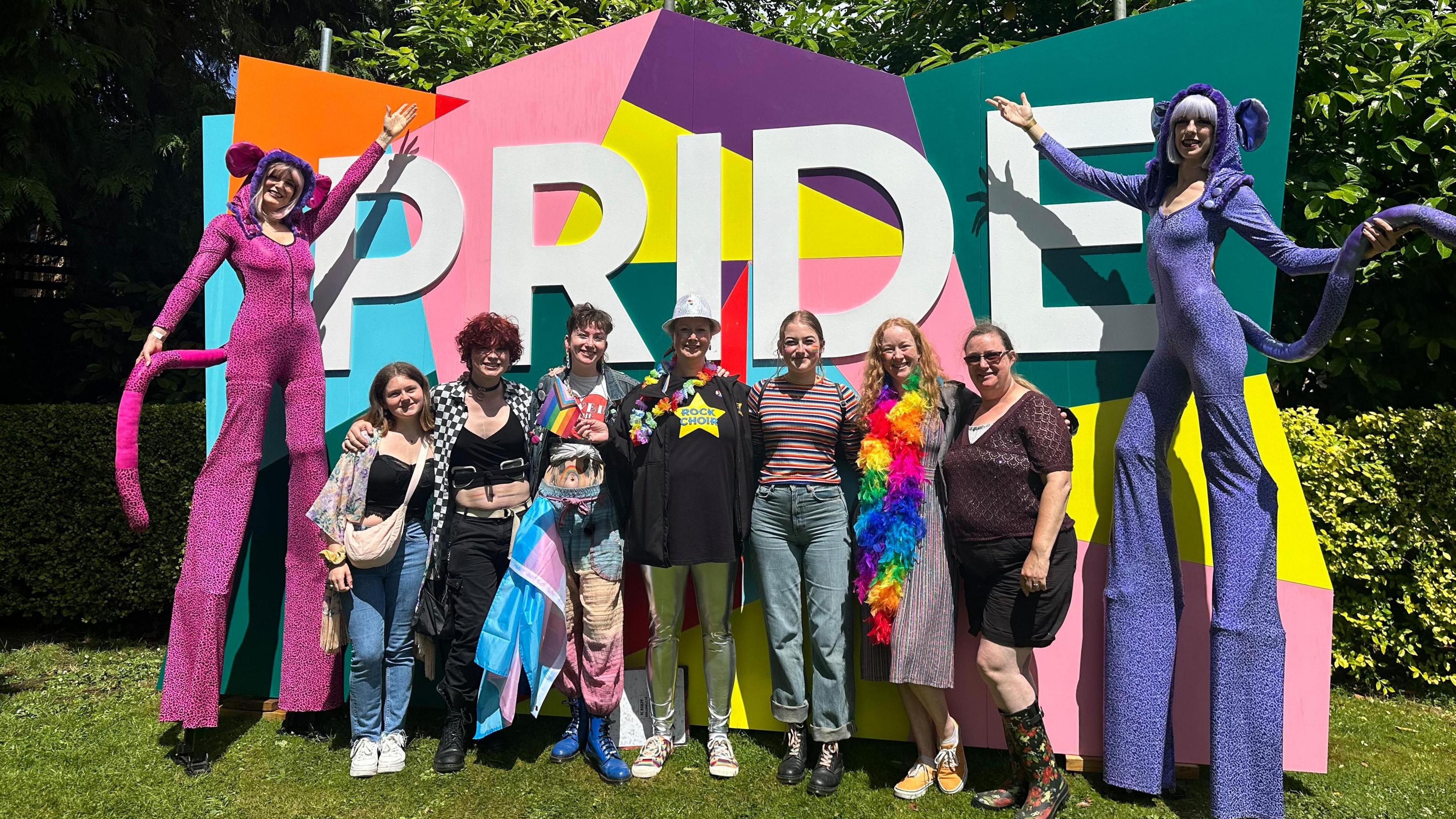 Seven friends, flanked by stilt walkers, pose in front of a large colourful Pride banner in a park