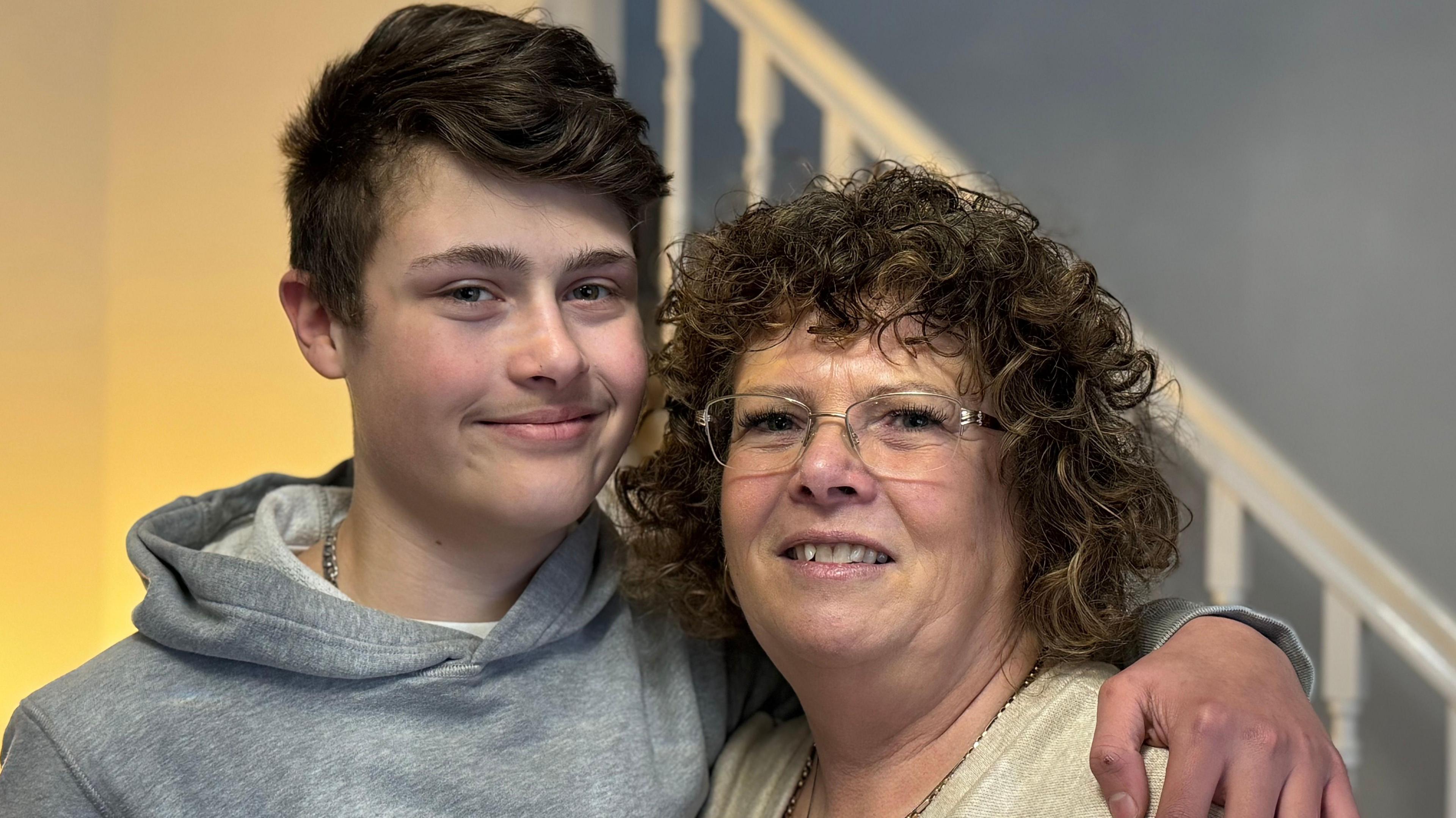 Jack Parton with his arm around his mum, Julie. Jack has a grey hoodie on, and Julie has a light yellow top on - she has curly, shoulder-length hair. They are both standing in front of a flight of stairs.