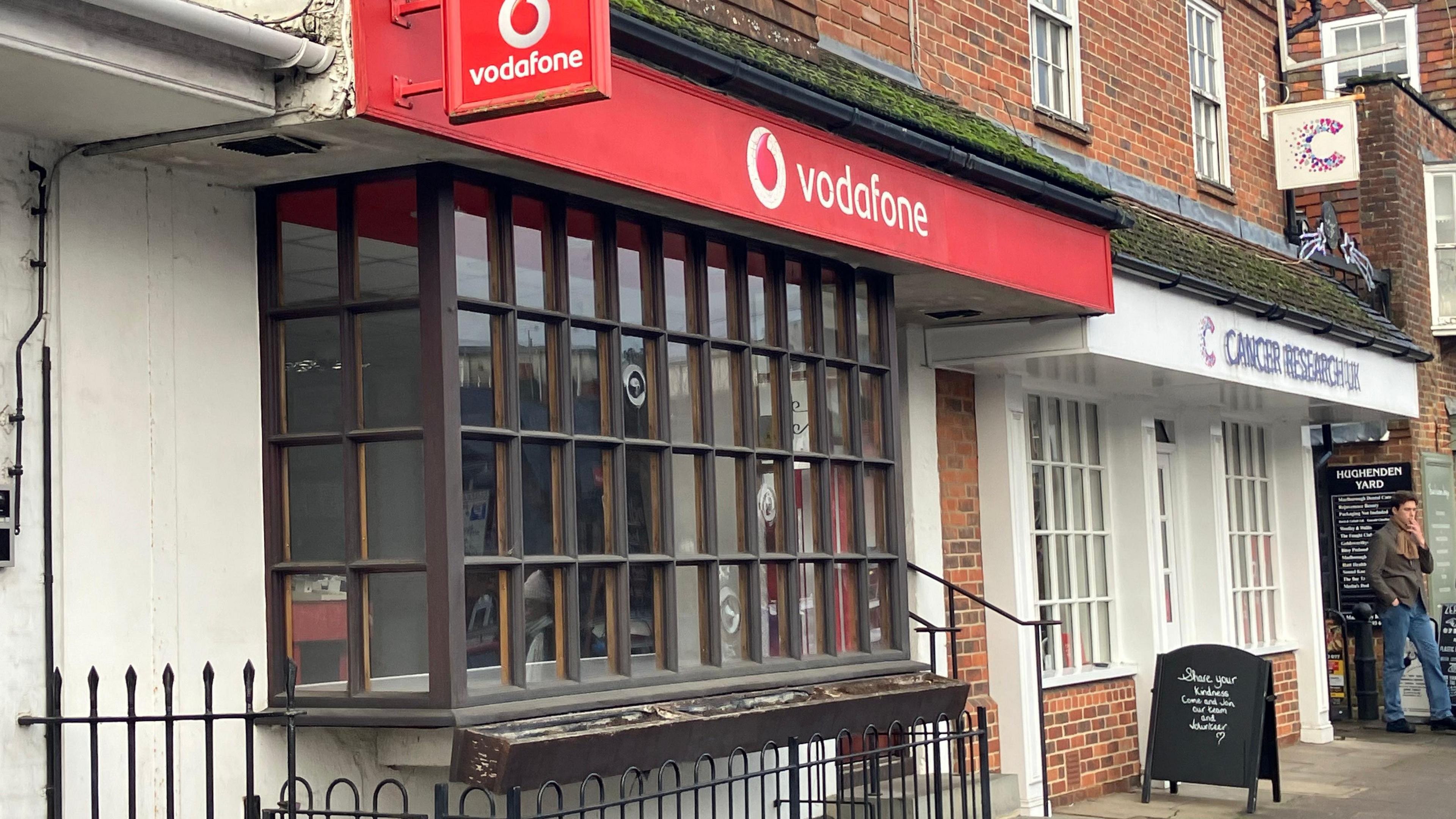 A view of the front of the Vodafone shop in Marlborough along the High Street. It is next to a Cancer Research shop. They are in red brick buildings.