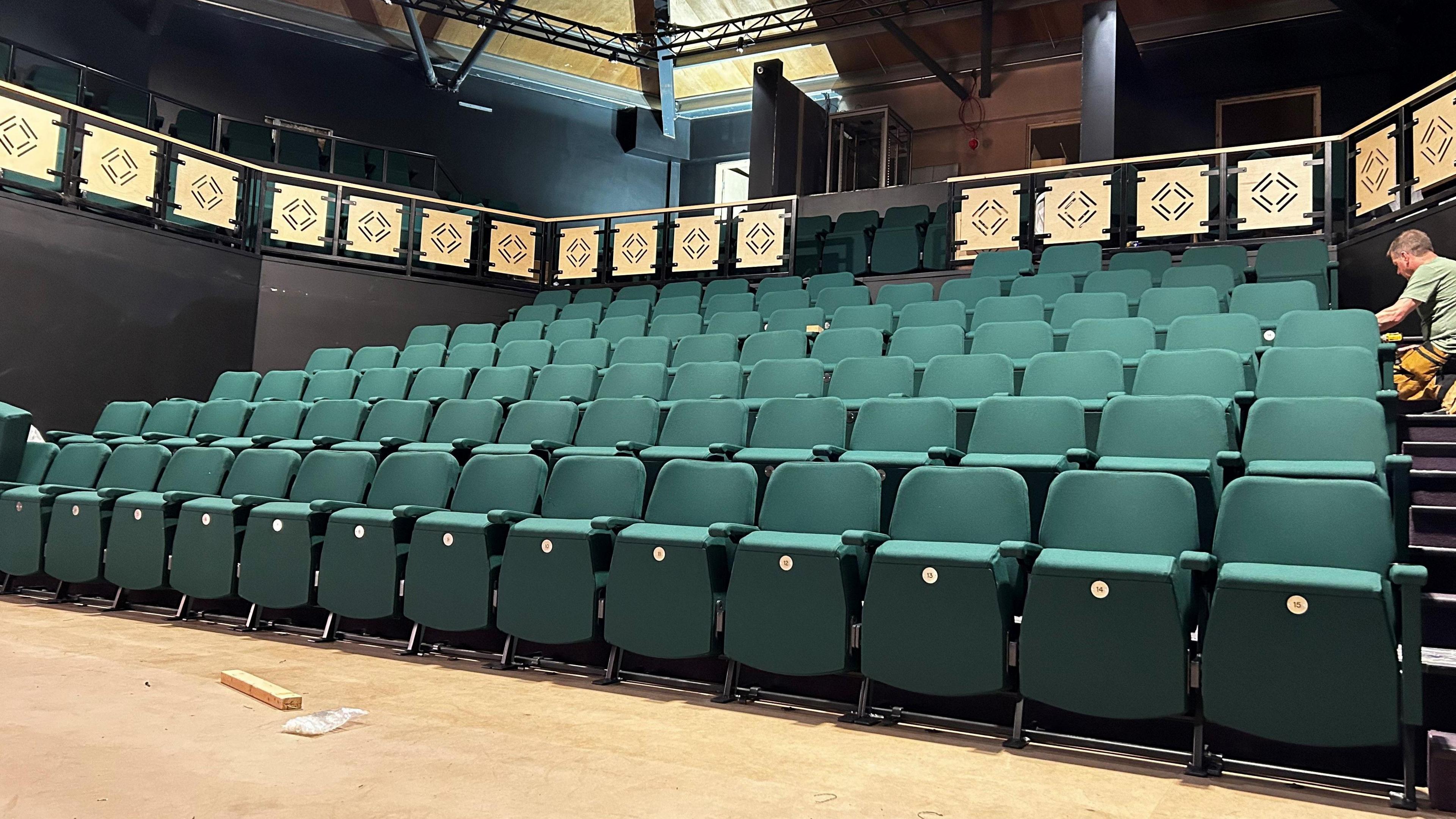 Seven rows of green seats in a theatre. Behind the seats is a barrier with golden panels on it with more green seats behind it. 