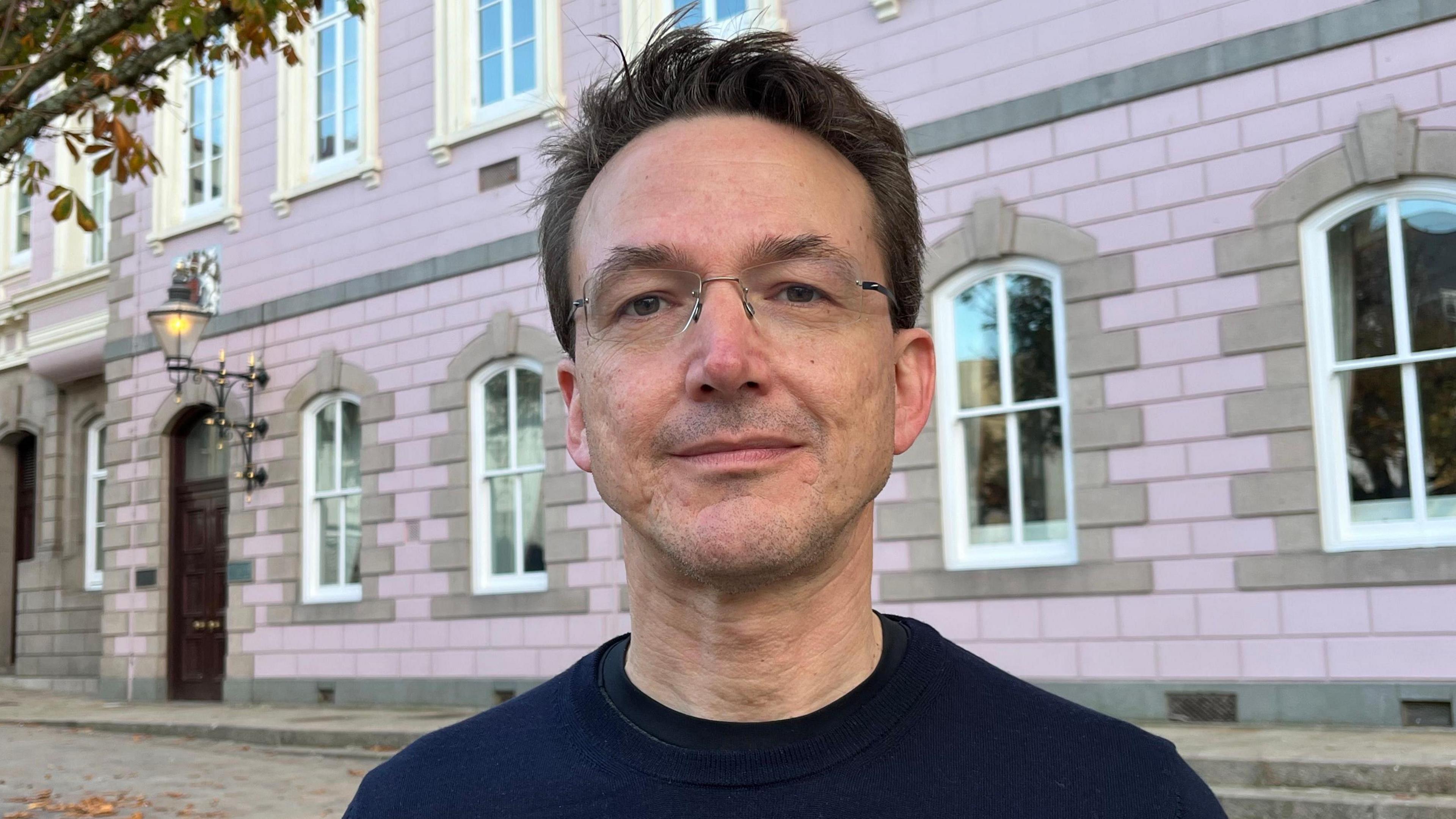 A shoulder shot photo of a man wearing a navy top wearing glasses standing in front of a pink and light brown government building.