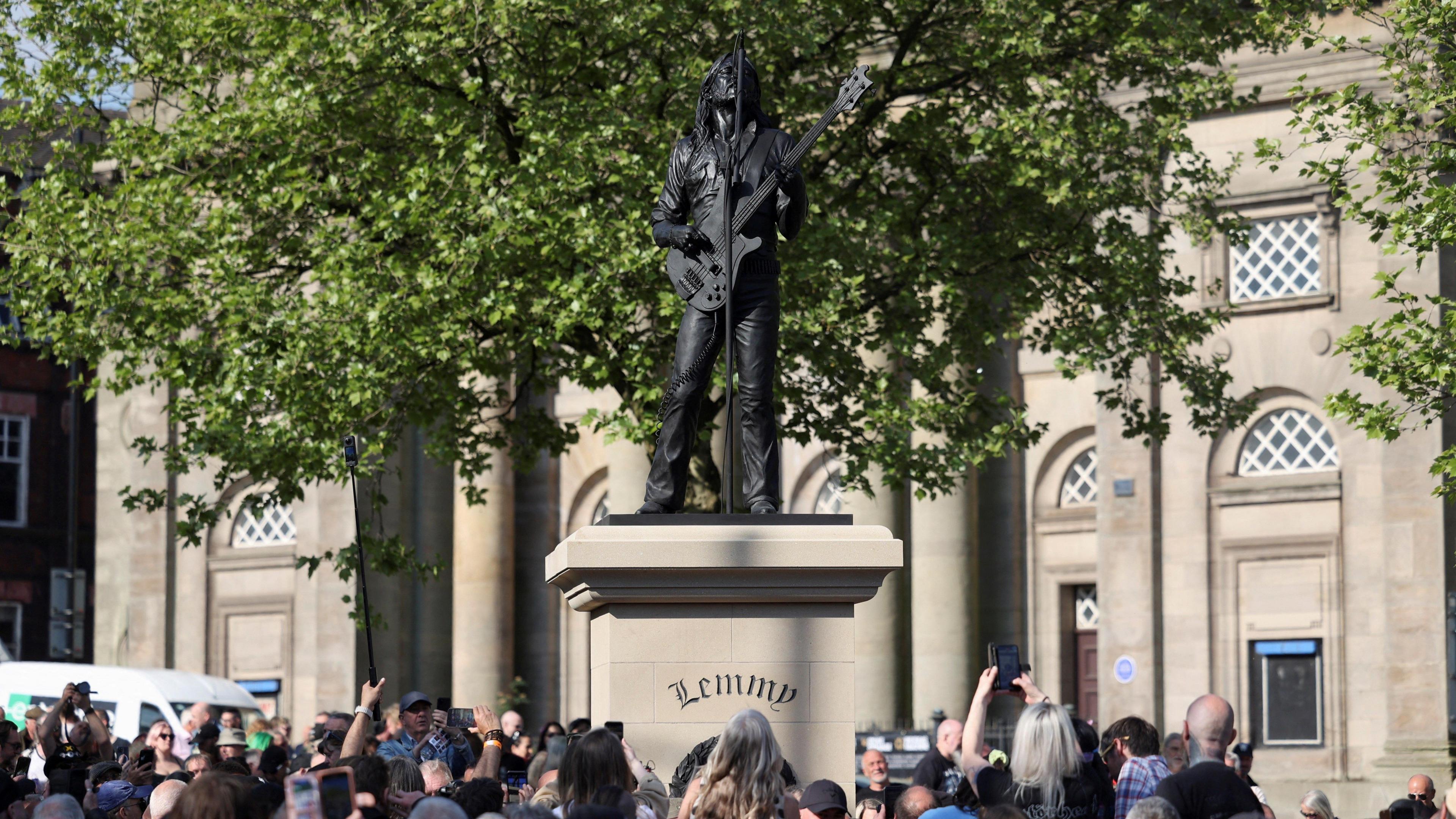 People gathered in front of the Lemmy statue in Burslem. Some are holding up their phones to take pictures. A large tree is behind the statue and a theatre building is visible behind it. 
