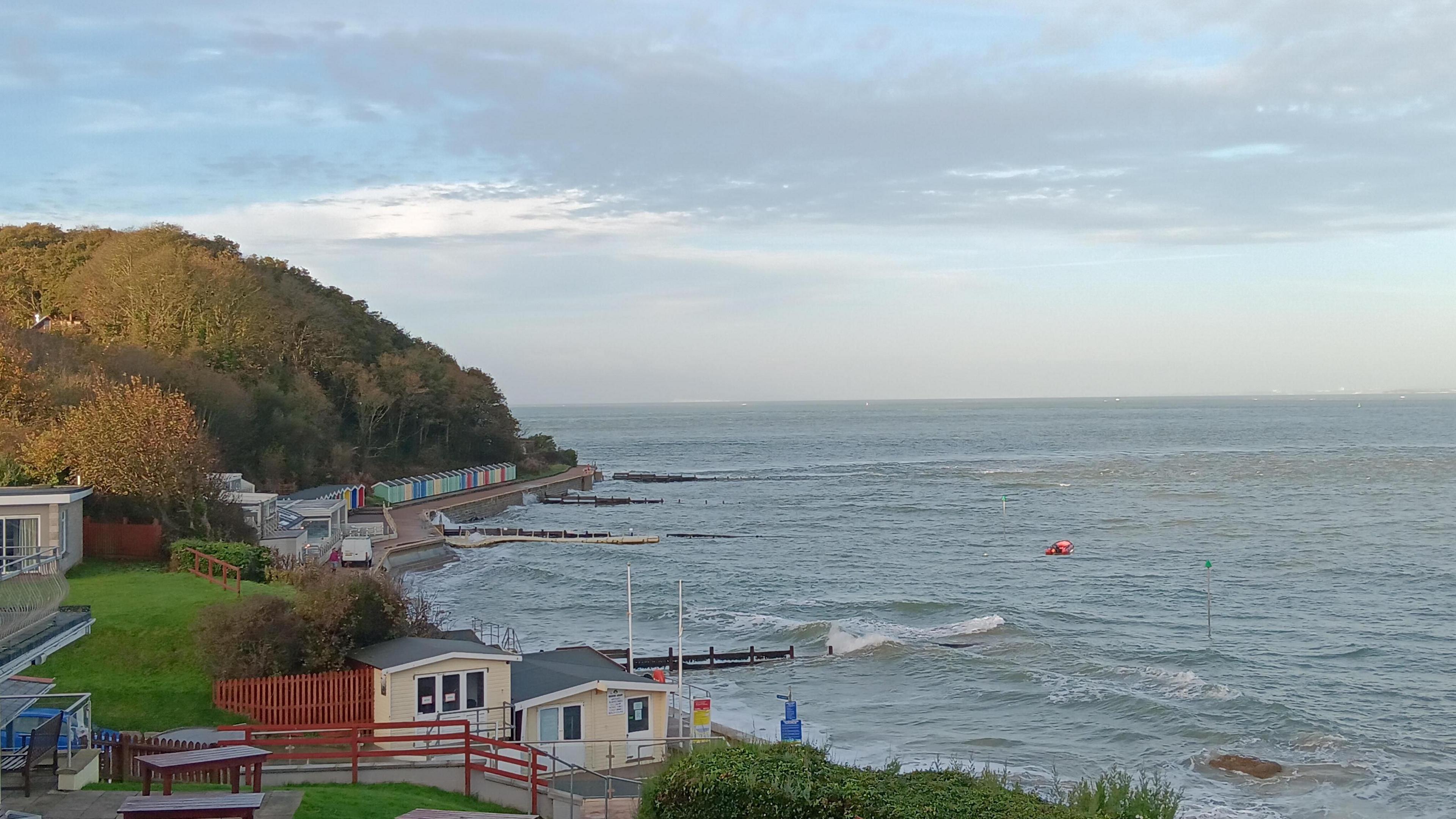 The tide is in at Totland Bay, with the water reaching the promenade lined with colourful beach huts and under a cloudy sky