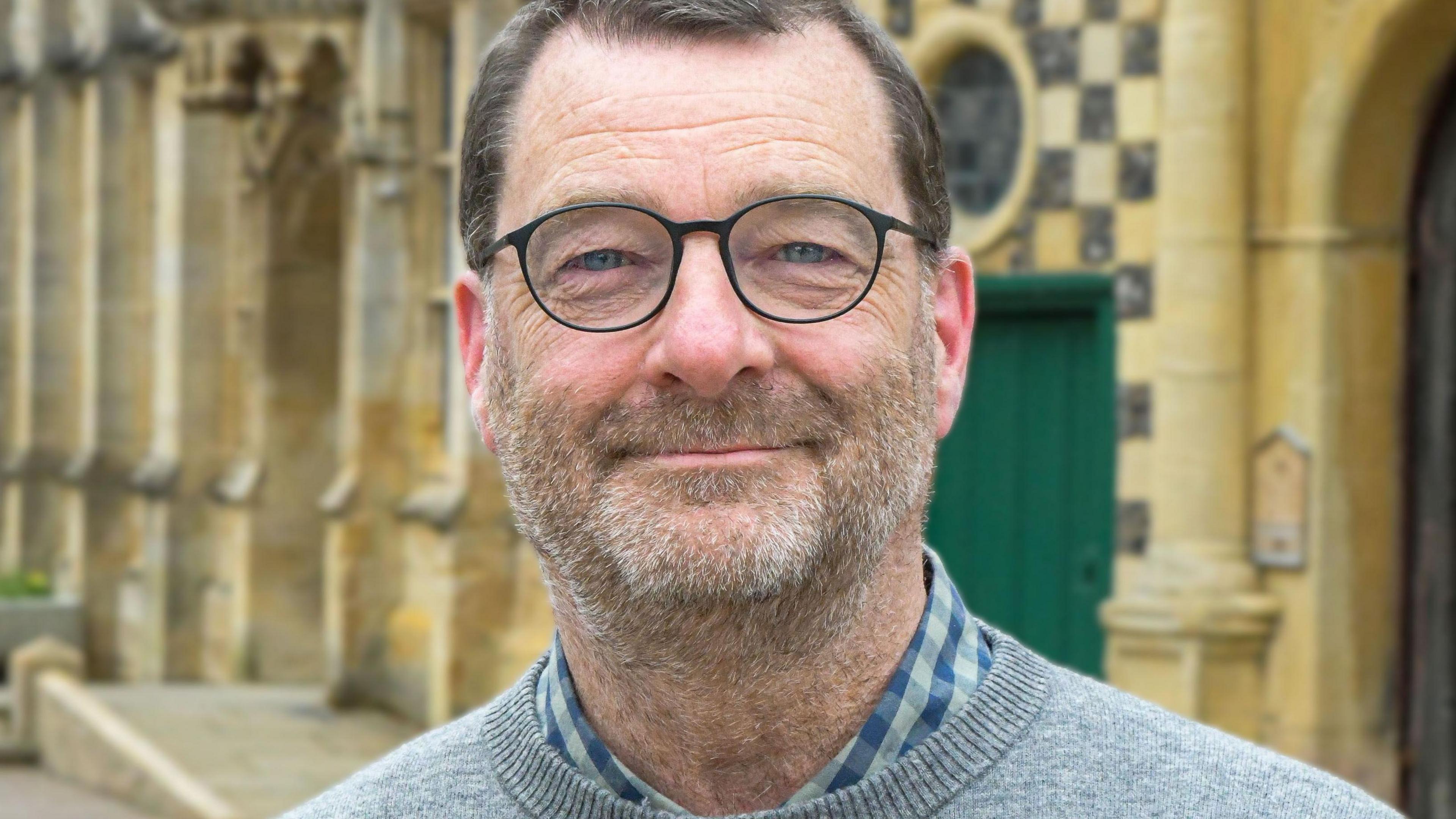 Simon Ring stands in front of the Guildhall. He is wearing a grey jumper. He has black glasses and a stubble beard. 