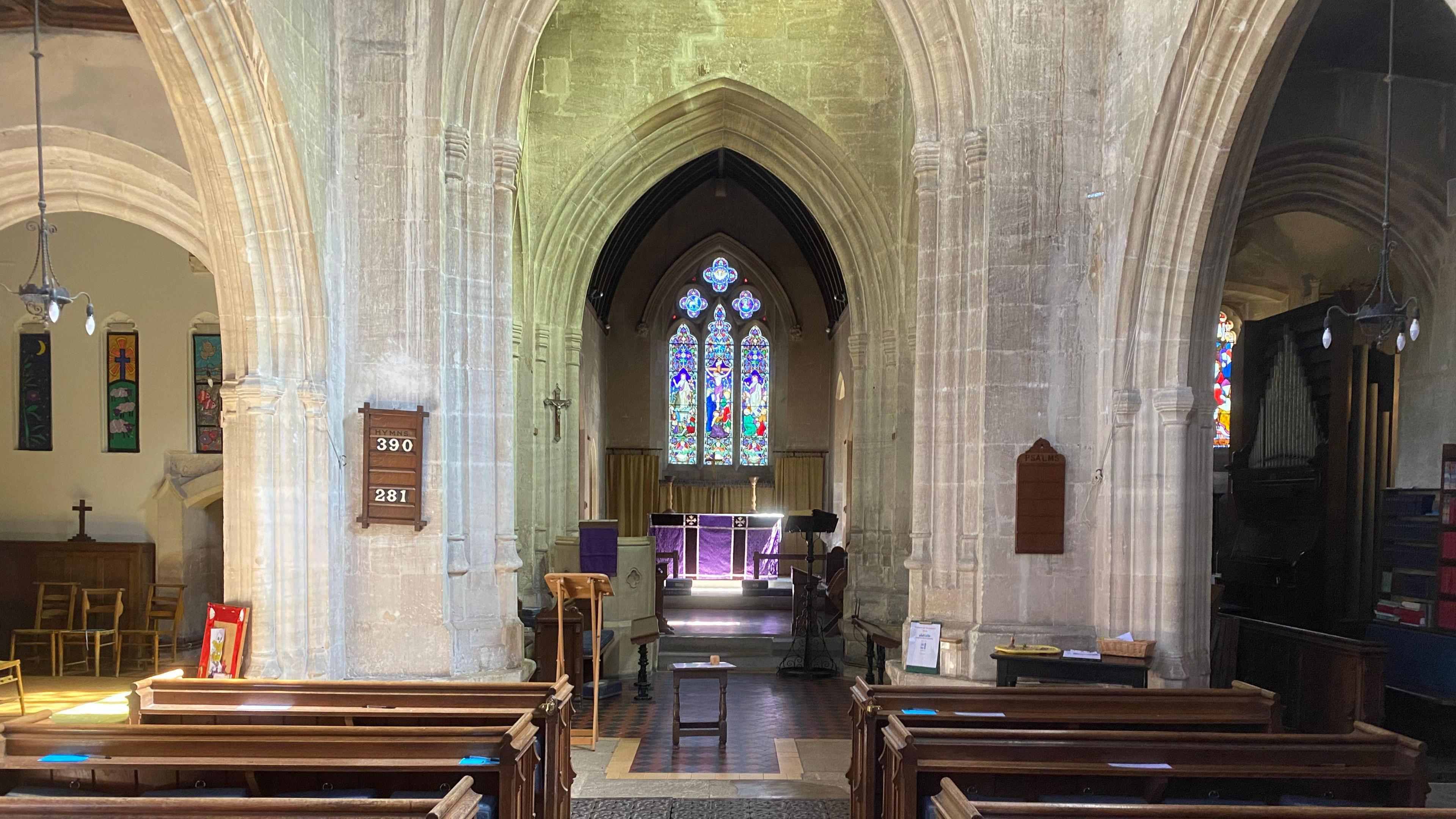 A view down a the central aisle of a church with a stained glass window at the end. Pews on either side with big stone arches. There are wooden noticeboards on either pillar and a lectern in the middle. Off to the left is a smaller chapel with a cross and hanging tapestries. On the right the organ can be seen in the distance.