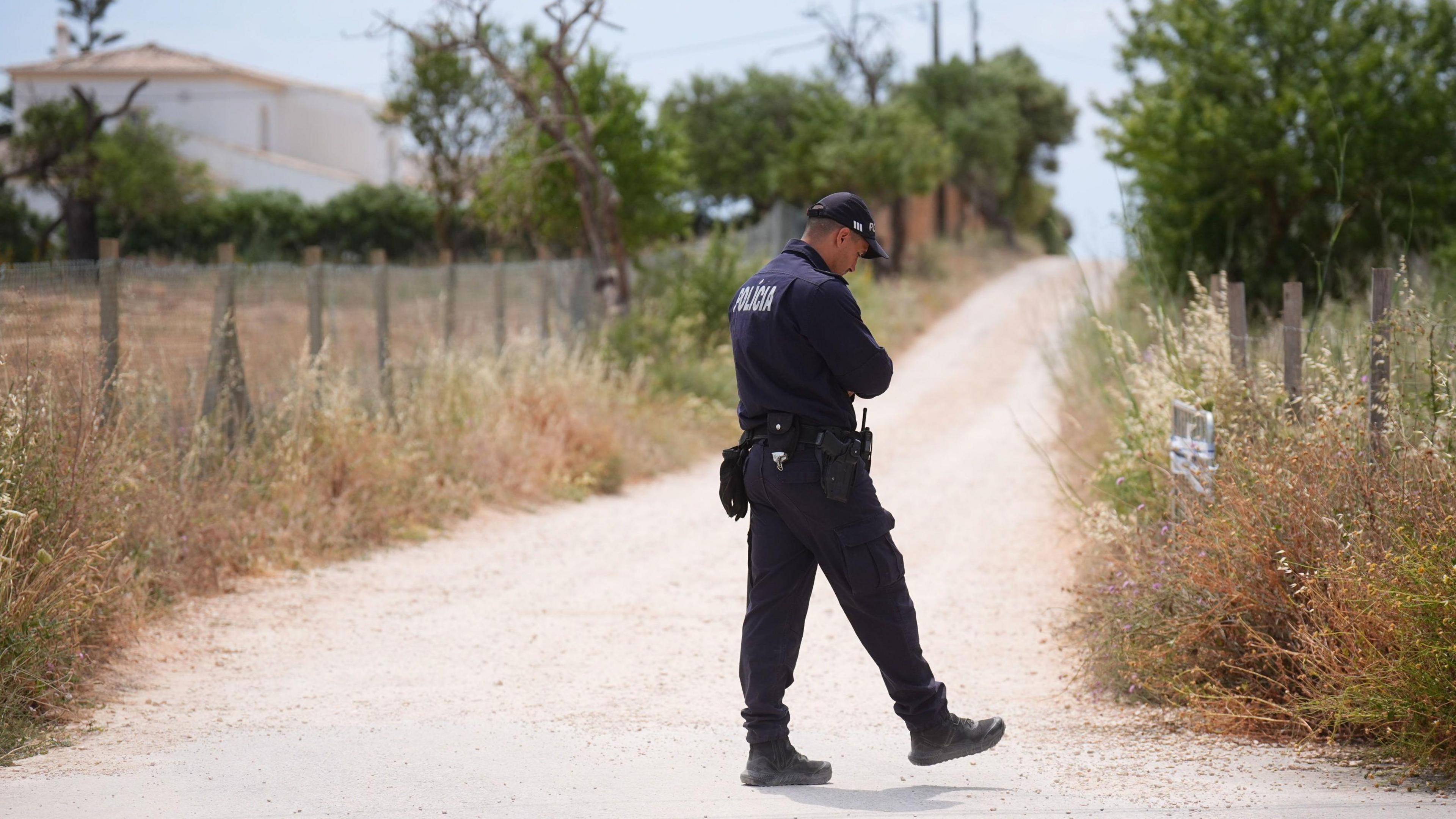 A police officer walks along a dry road with tall grasses either side. A white house is in the distance.