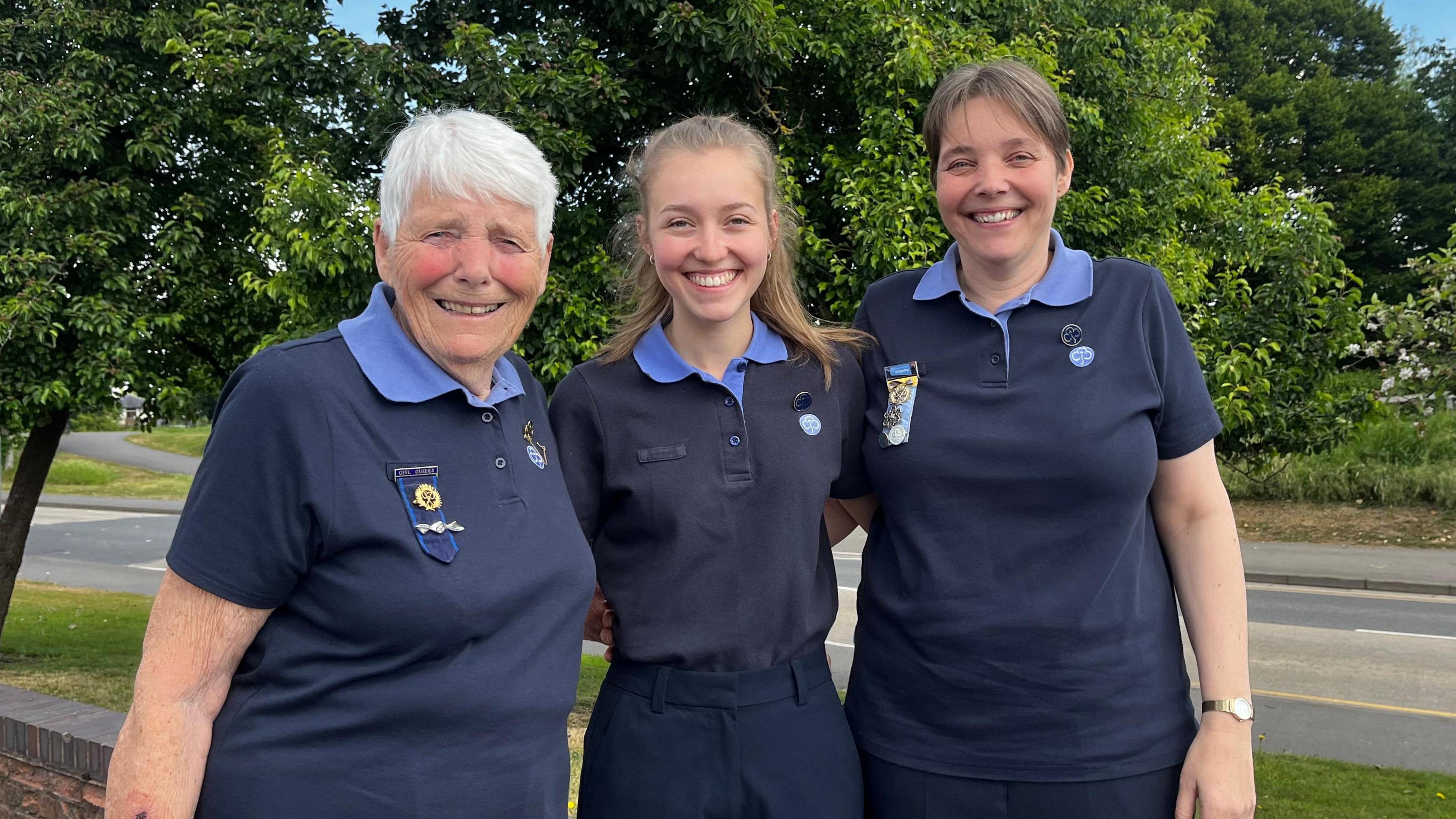 (L-R) Gillian Watson, Megan Megahey and Hazel Megahey. They are standing outside, wearing Girlguiding uniforms. They have their arms around each other and they are all smiling. There is grass and trees in the background and it is sunny.