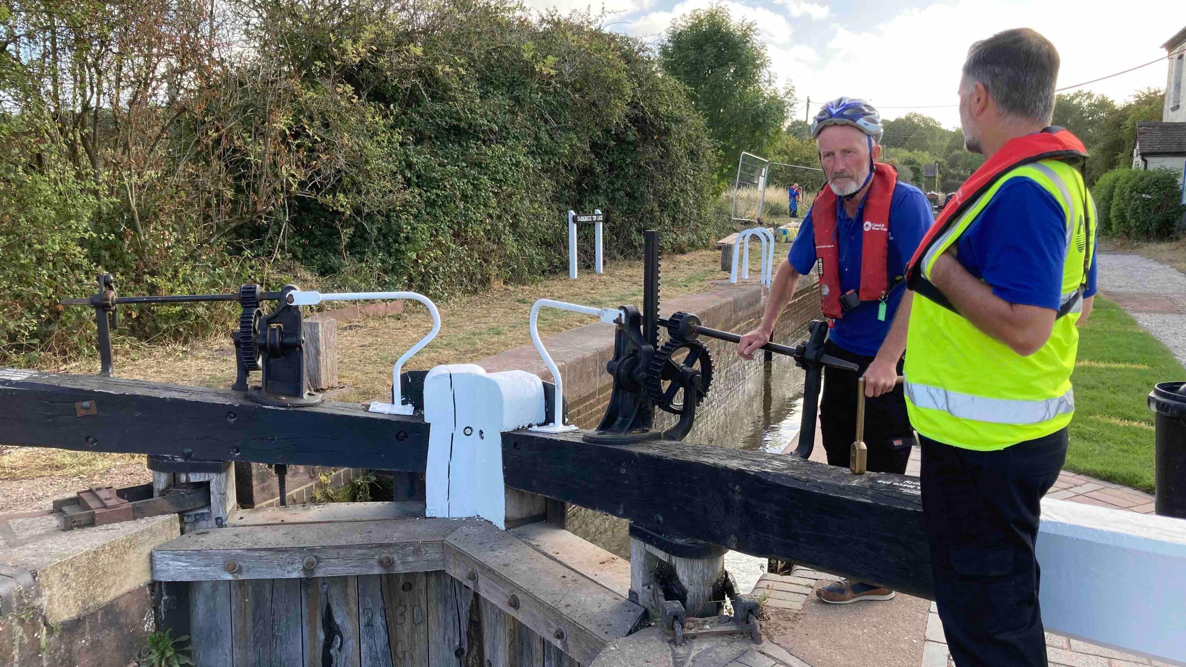 Two workers from the Canal and River Trust wearing blue polo tops and life jackets stand next to the wooden top lock. Lock is made up of a wooden structure blocking the water and two large wooden bars that are used to open it on the surface.