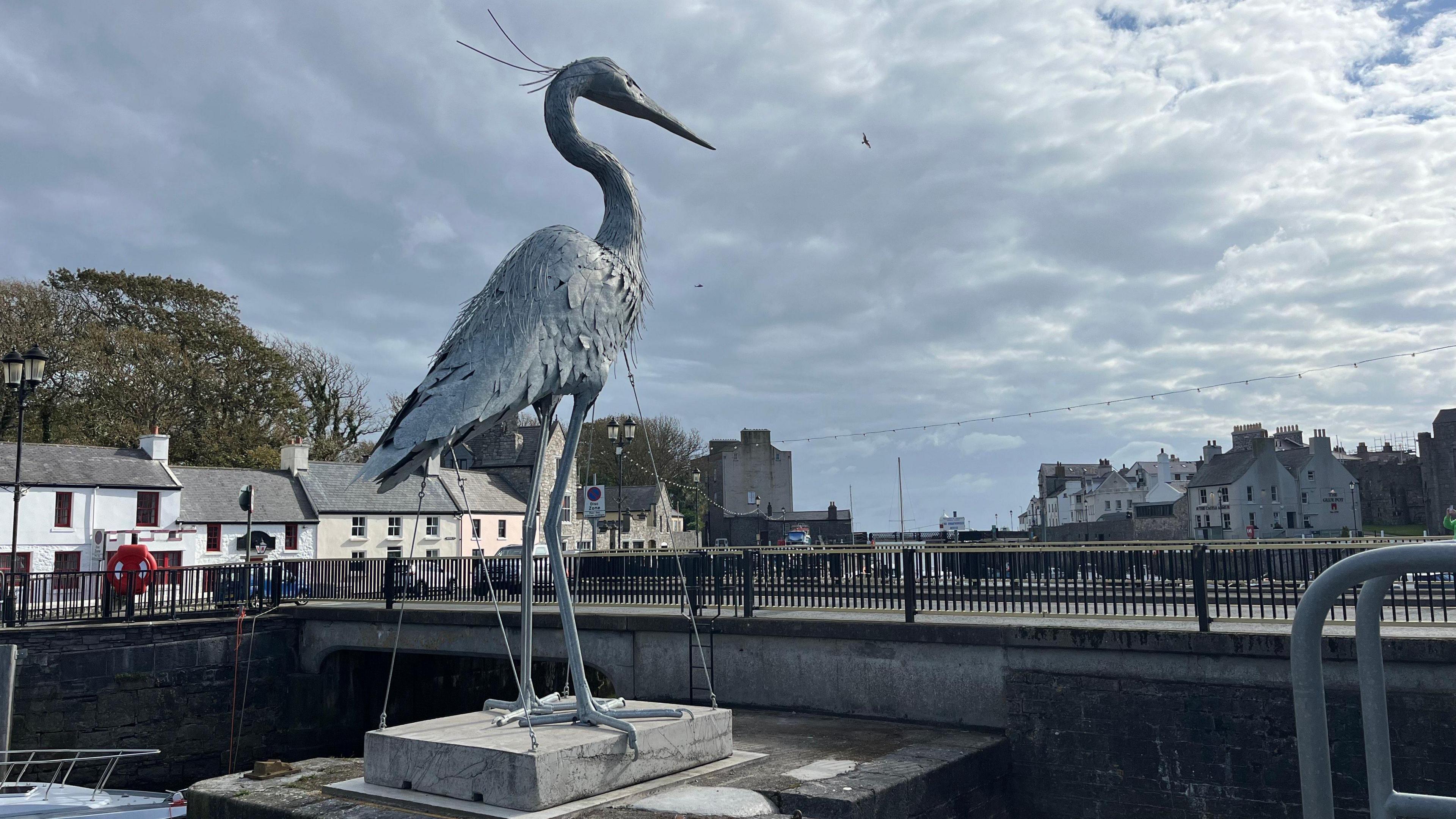 A giant silver heron sculpture next to a road bridge over Castletown Harbour with buildings in the background, the heron is taller than the buildings.