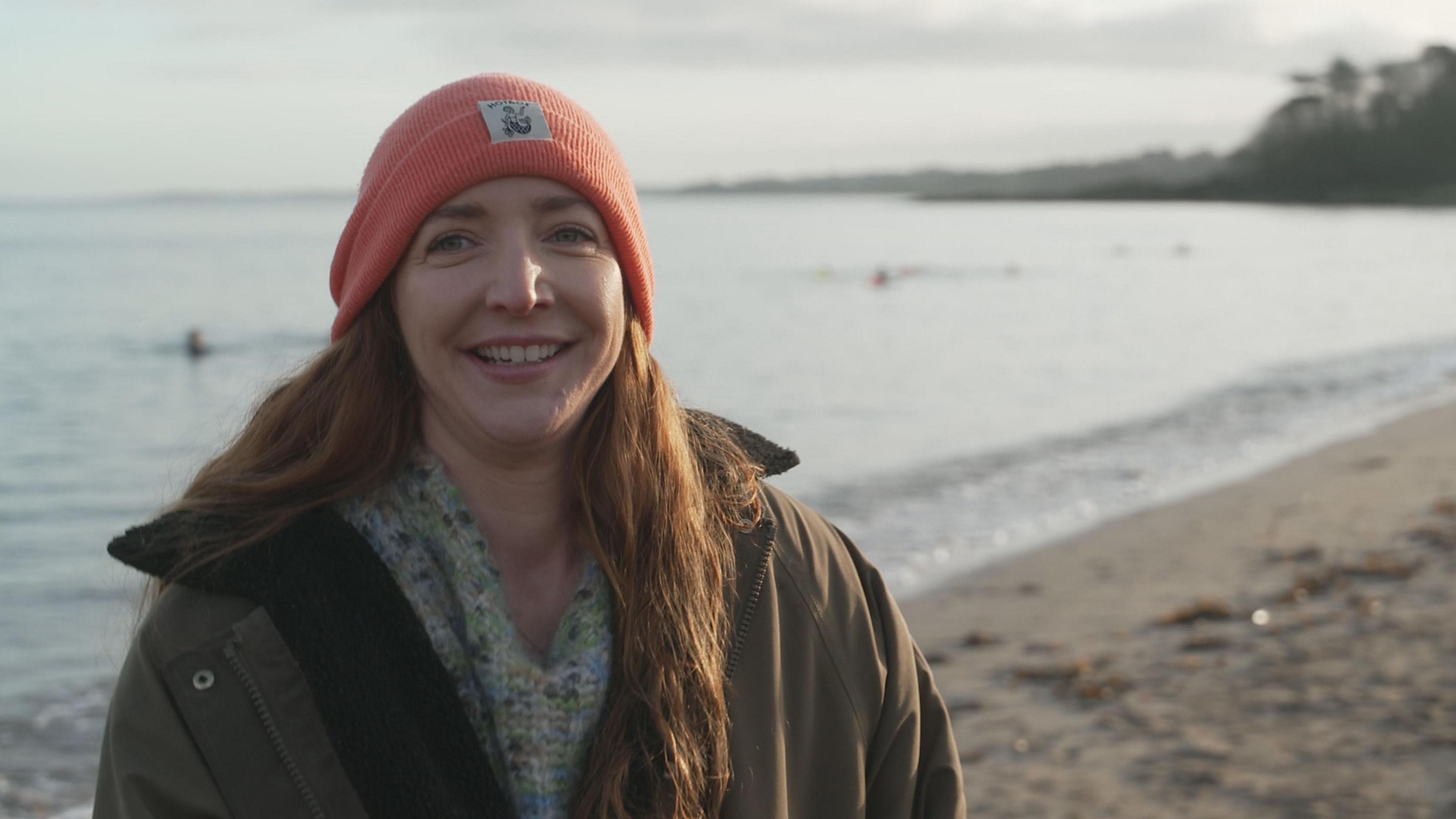 Helen is standing by the water at Helen's Bay beach with some swimmers out of focus behind her. She has long, wavy, ginger hair which hangs over her left shoulder. She smiles, wearing an orange woolen hat, a woolen scarf with green, cream and blue material, and a green coat. There is a hint of an orange sunrise to her right hand side. 