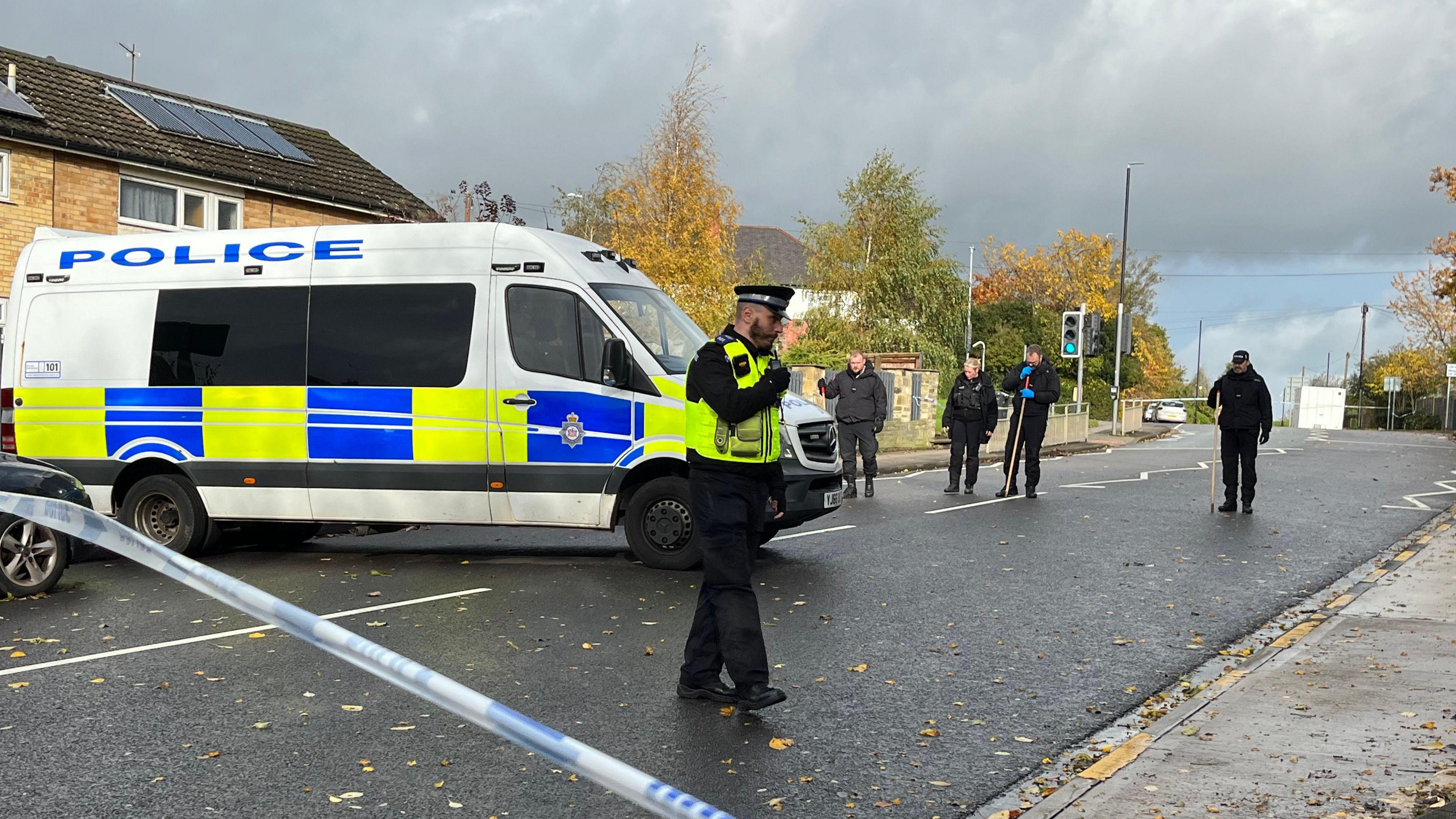 A police van is parked across a residential street. Several officers can be seen staring at the floor as part of a search.