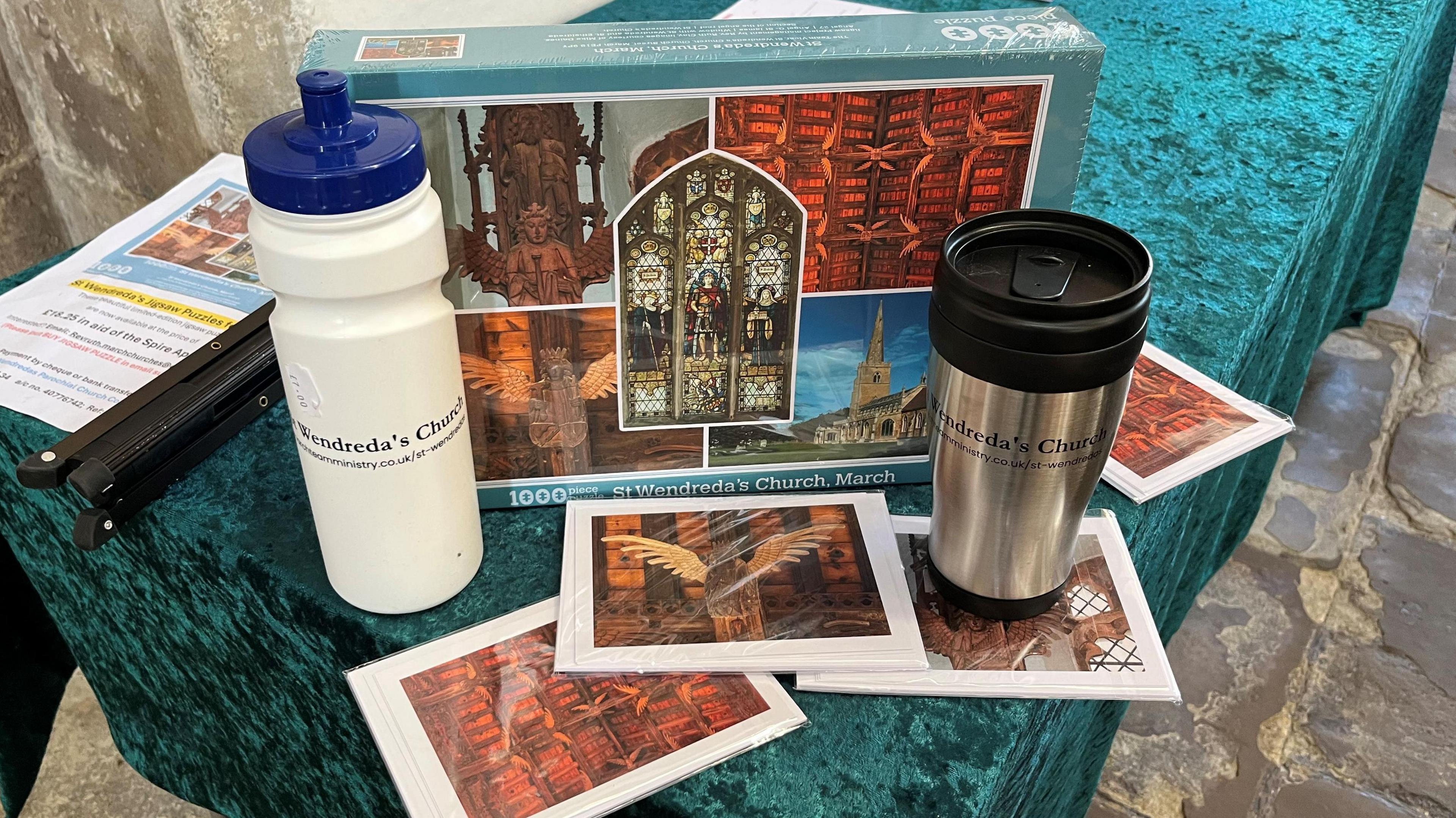 A selection of merchandise on a trestle table inside the church, including a 1000-piece jigsaw puzzle of St Wendreda's, and some water bottles and postcards bearing its image.