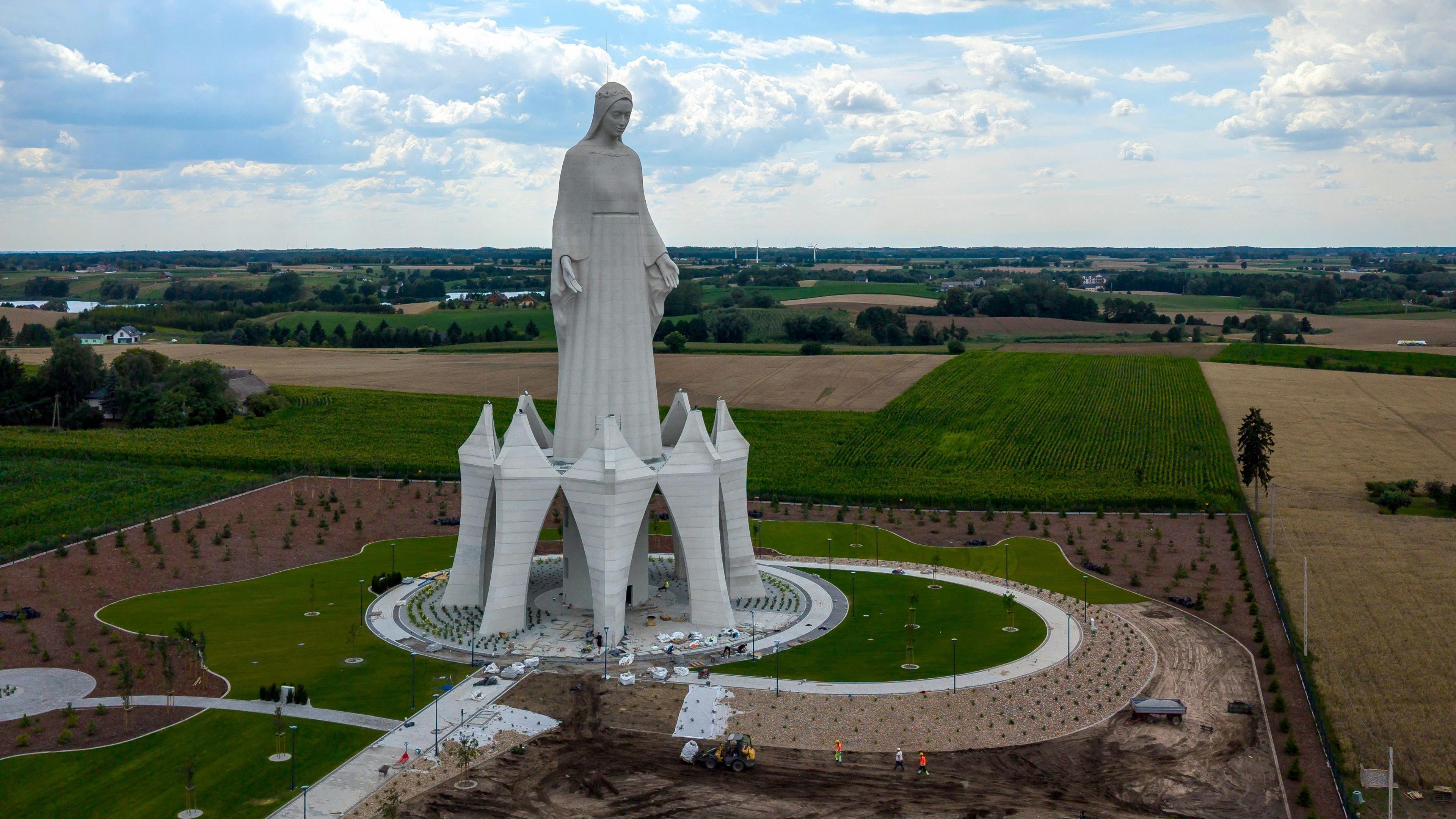 A statue of the Virgin Mary rises out of green and yellow fields in Poland on a cloudy sky. 