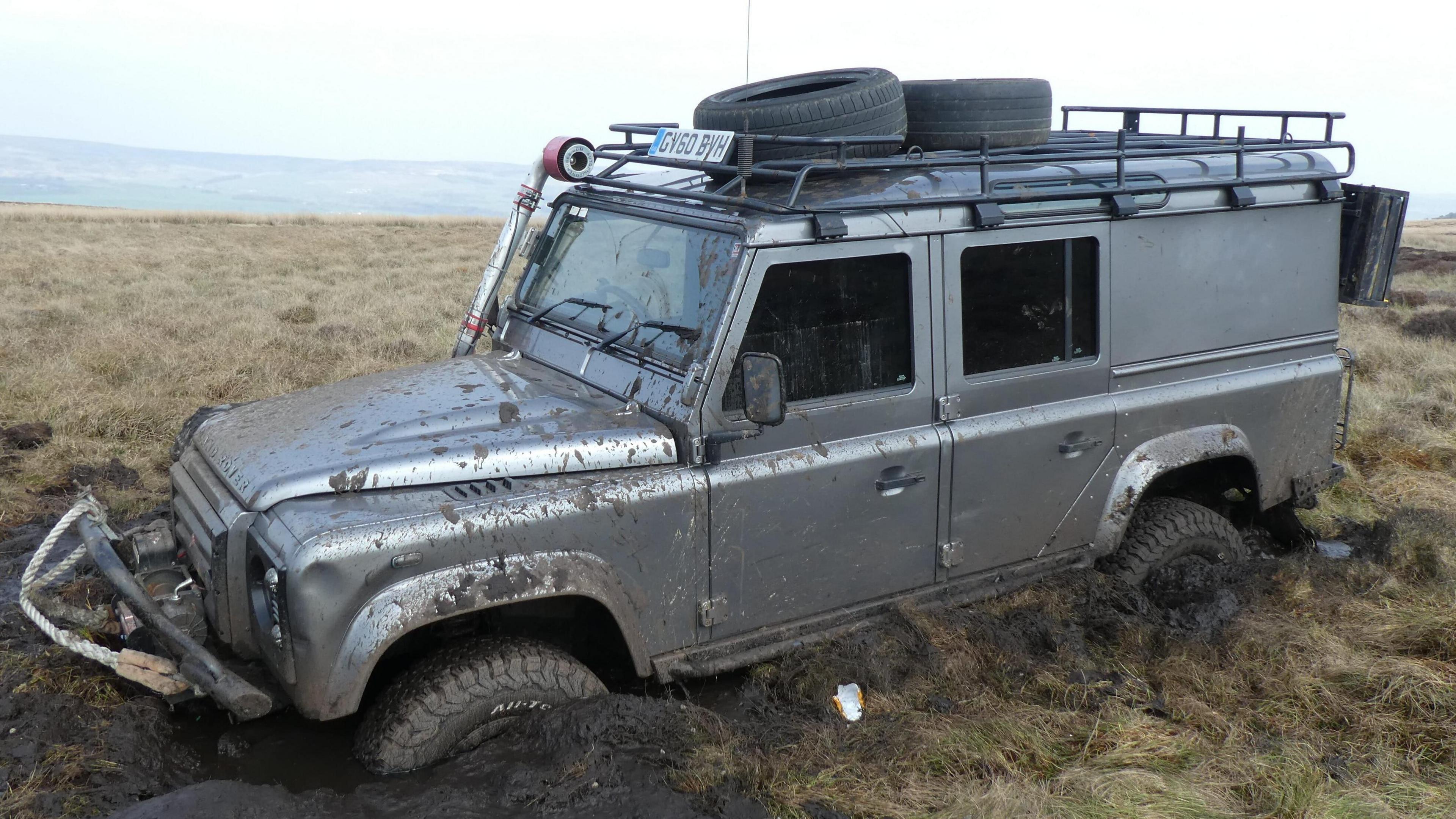 Picture shows a grey-silver coloured jeep stuck in mud in a field. Its wheels are almost completely submerged in the mud. The wider Ilkley moorlands and rolling hills can be seen in the background.