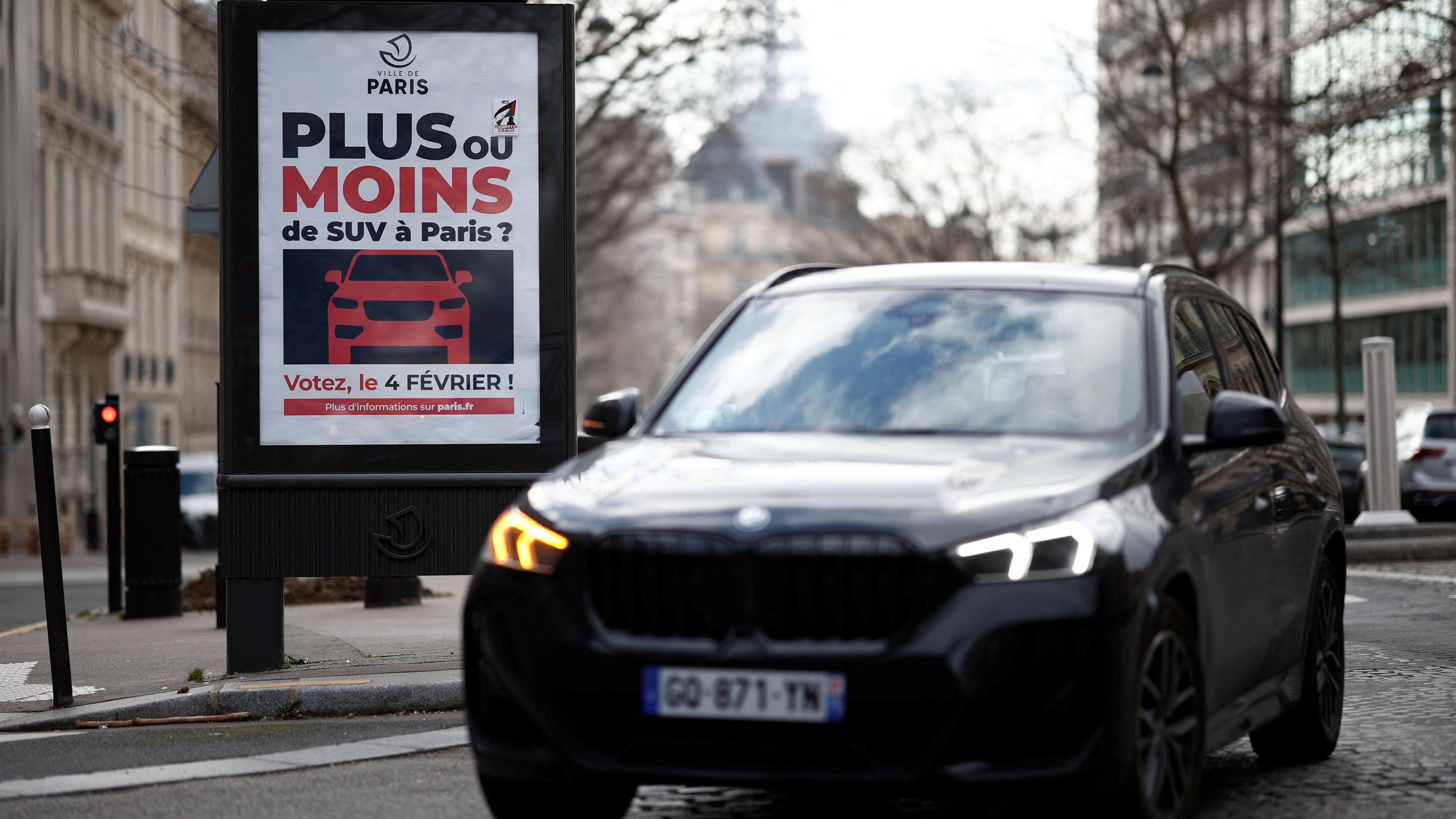 A large black car passes in front of a poster